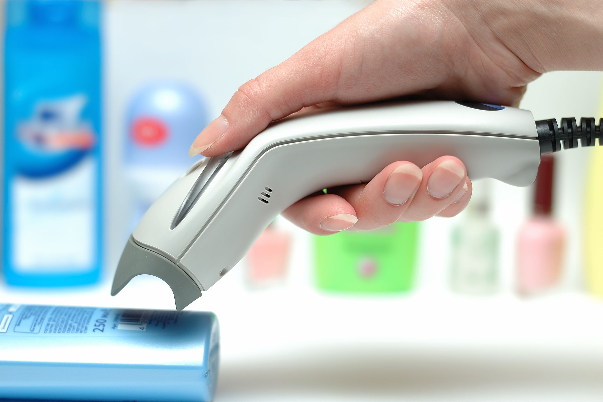 Closeup side-view of female hand holding a scanner, scanning a barcode of the back of a bottle of shampoo on a white countertop, a row of personal care items and nail polish is blurred in the background
