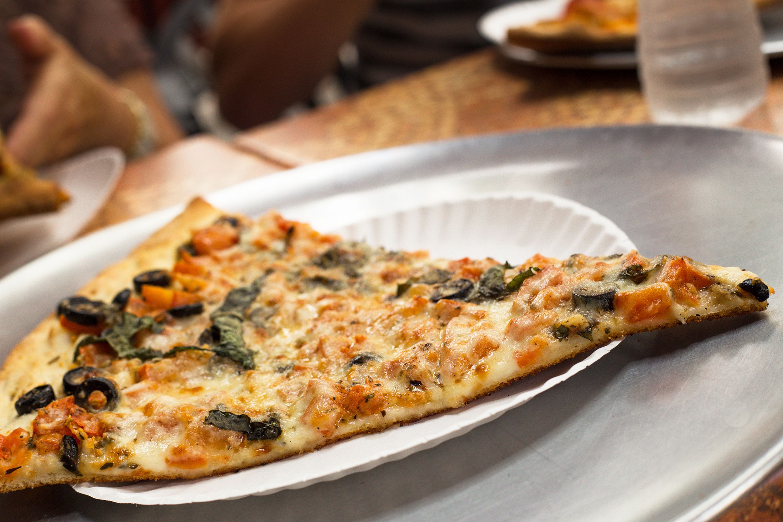 Closeup of slice of traditional New York style pizza on a paper plate on a stainless steel round serving tray, cheese, chicken, and black olives, blurred background of table, seats, and people eating