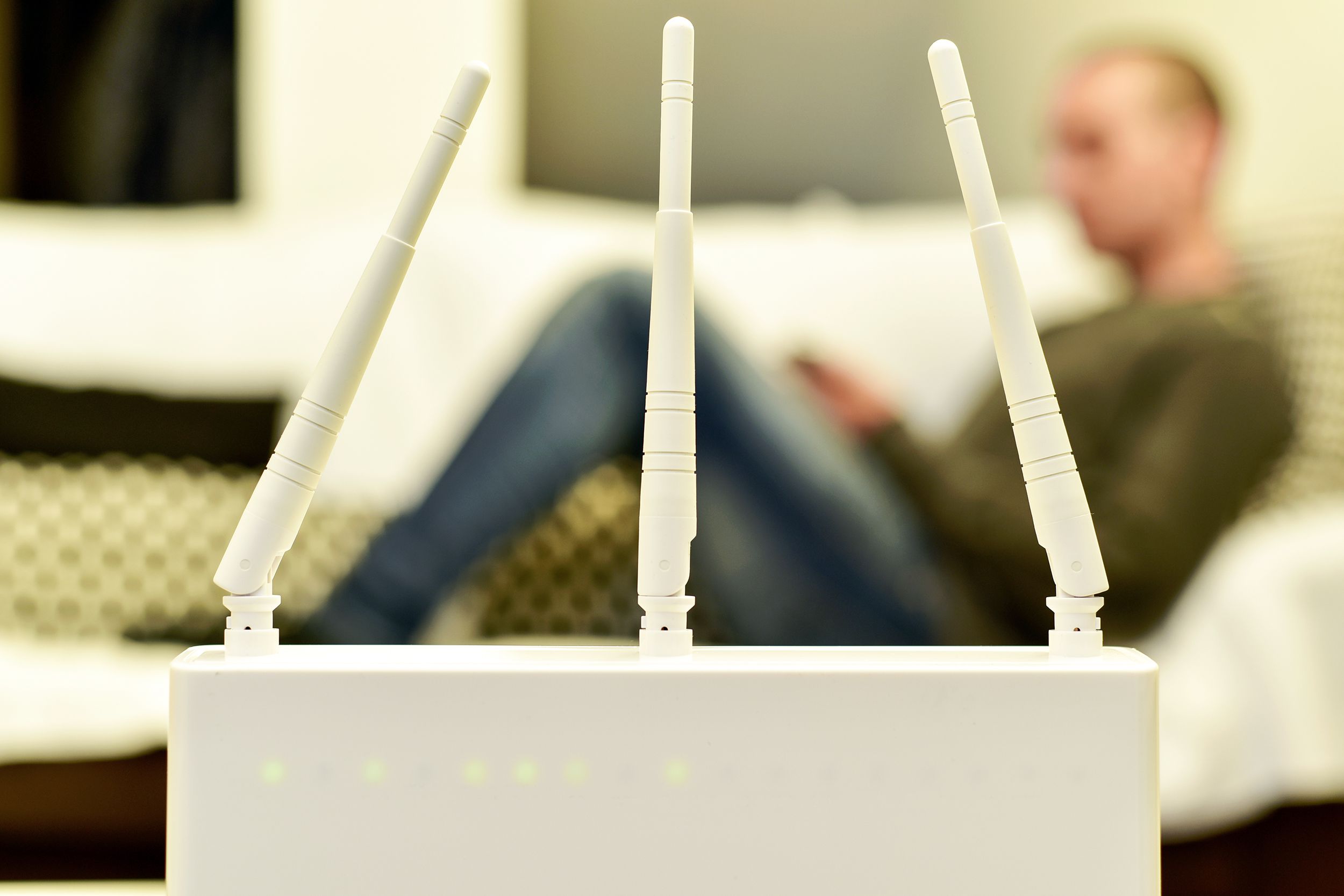 Closeup of three antenna on a white wireless router, selective focus, man is using a smartphone while lying on a couch with two large windows, dark at night, blurred in the background