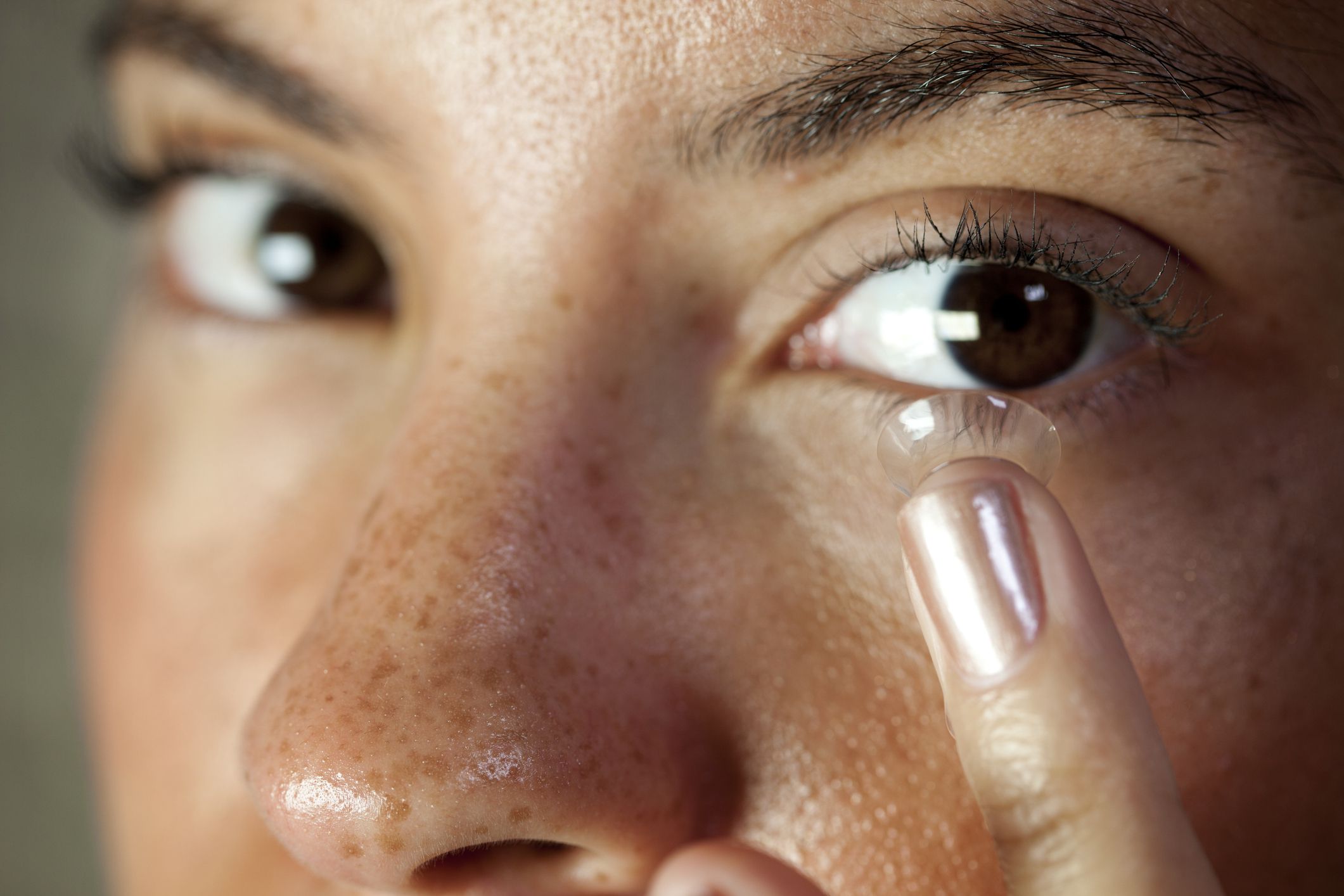 Closeup of woman about to insert a contact lens into her left eye, selective focus, both eyes looking slightly to the right side, her right side of face blurred