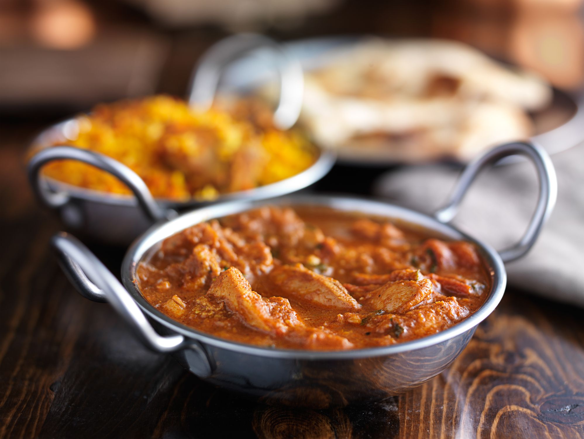 Closeup of curry in a metal serving dish at an Indian restaurant with another curry in a metal serving dish and naan with a blurred background