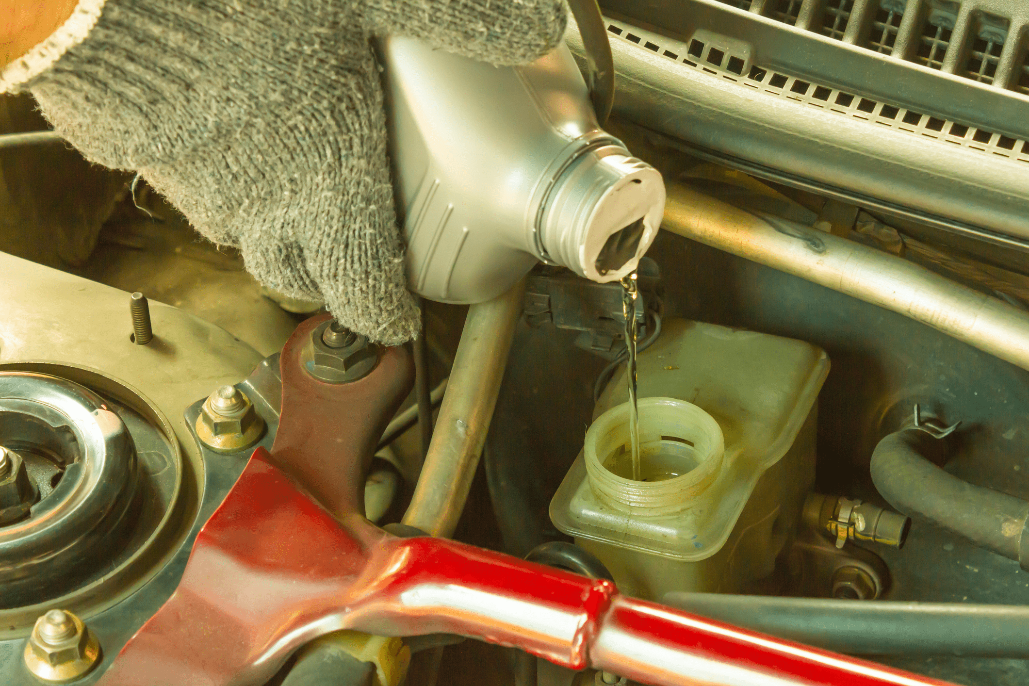 Closeup of two grey gloved hands pouring new brake fluid into a car engine, car engine in the background