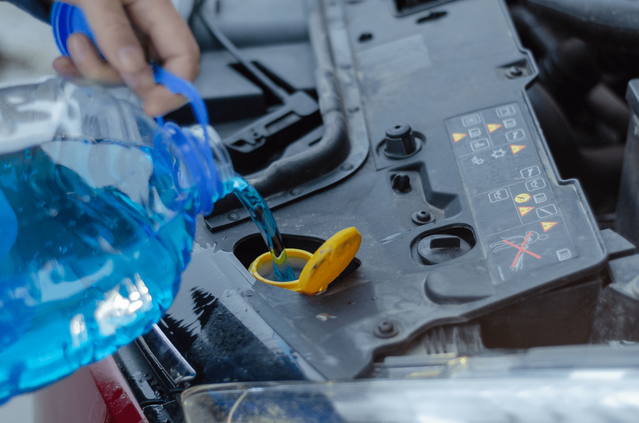 Closeup of windshield wiper fluid in a gallon jug being poured under the hood into a car, area is indicated by a yellow cap, car engine is blurred in the background