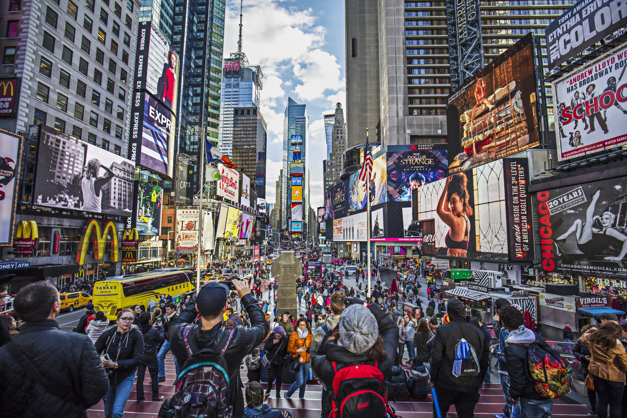 Crowds of people in the center of Times Square, Manhattan, large advertisements on buildings on both sides, dramatic clouds and blue sky in the background