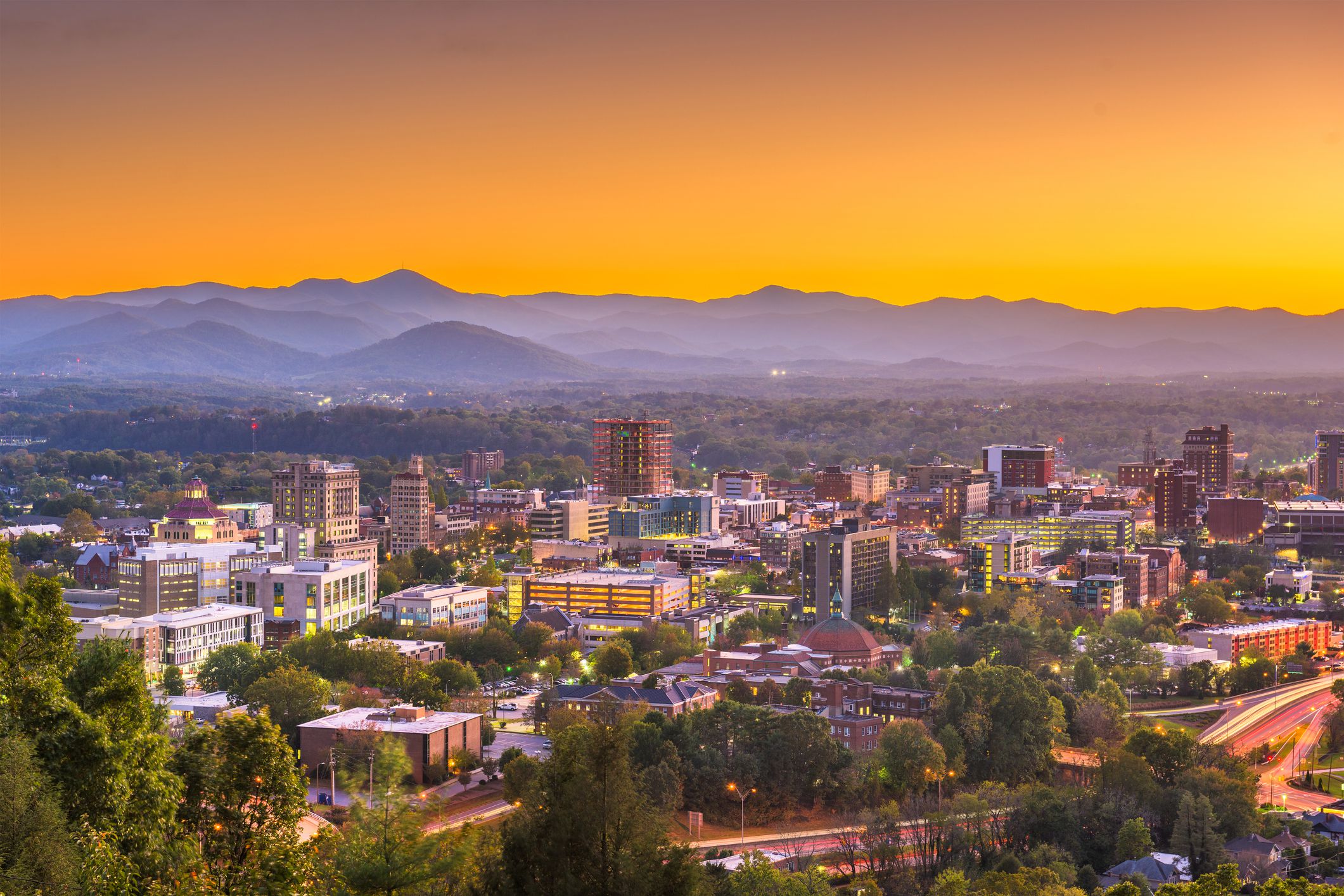 Downtown skyline of Asheville, North Carolina, during dawn, trees in the foreground, on a hill, blue mountains in the background with a dramatic orange sky