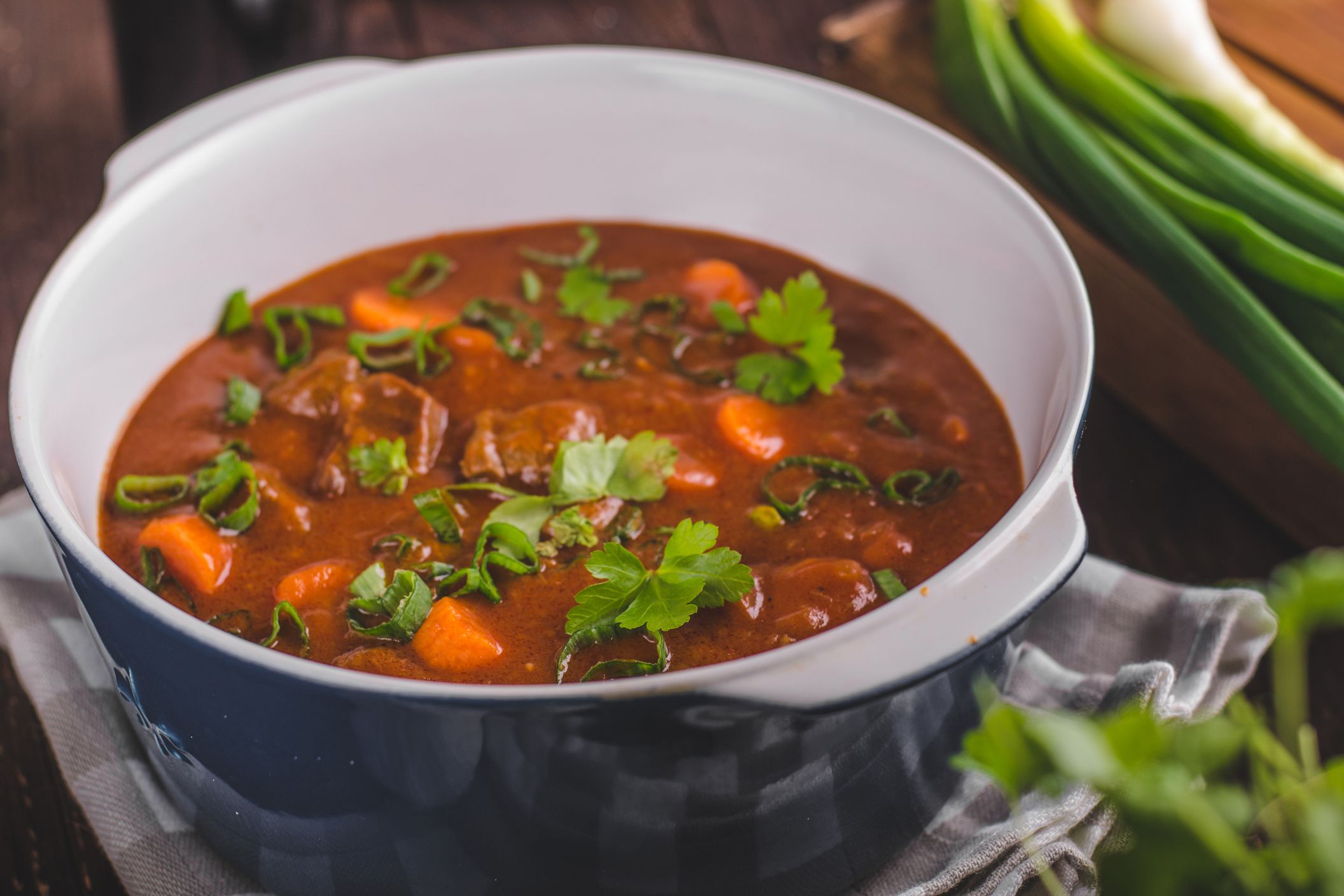 Easy 5-ingredient beef stew in a blue with white inside ceramic bowl on a matching blue checkered napkin with a blurred background of ingredients and wooden table