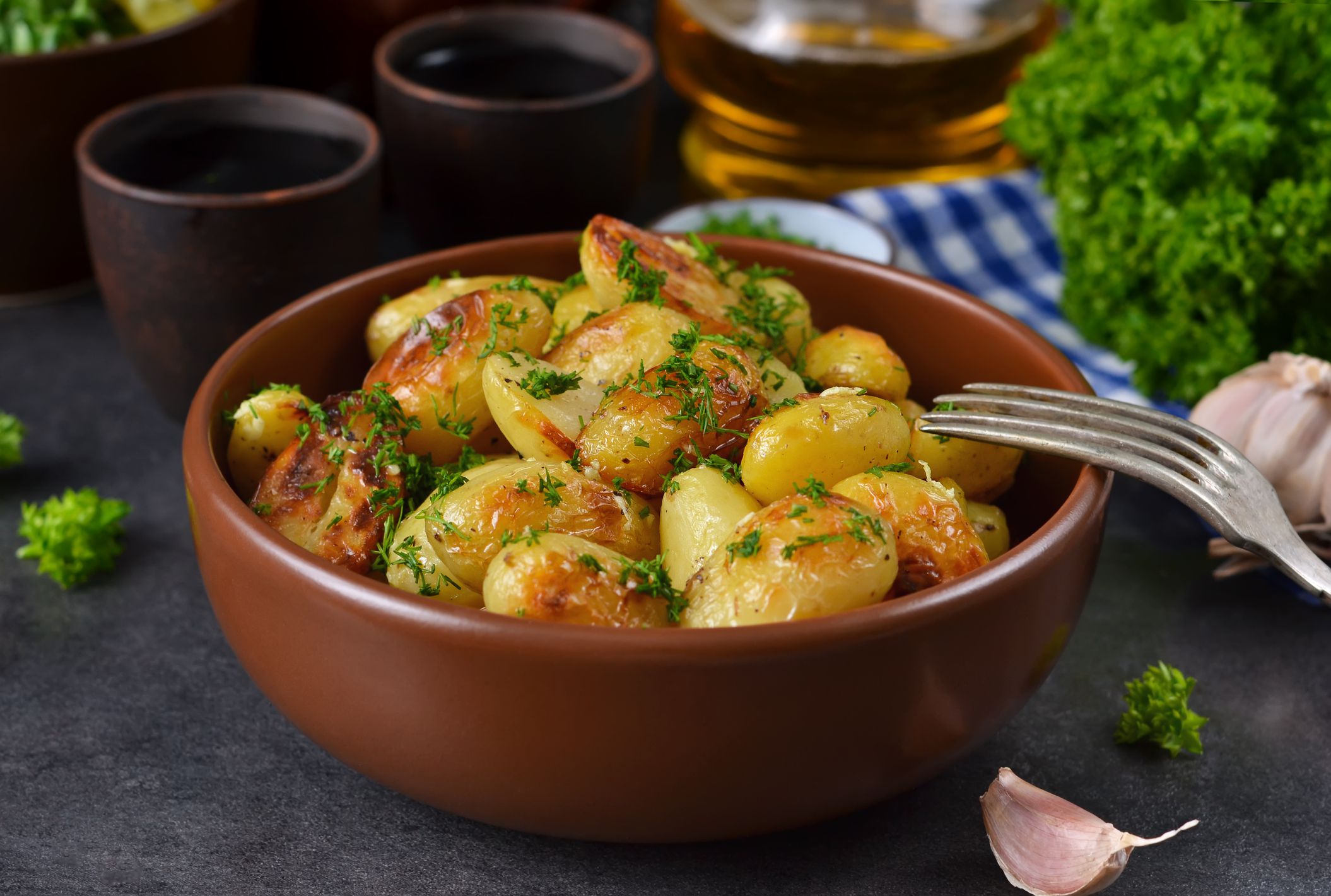 Easy grilled potatoes in foil with garlic in a terracotta bowl and fork placed on rim of bowl with two terracotta cups of tea, garlic, a bottle of oil, and more in the background