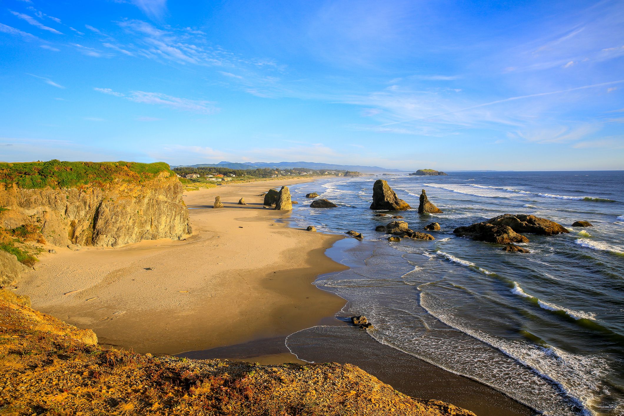 Expansive view of Bandon Beach, Bandon, Oregon, cliffs line one side of sand with many large rocks in the ocean with waves, a few wispy clouds in a blue sky