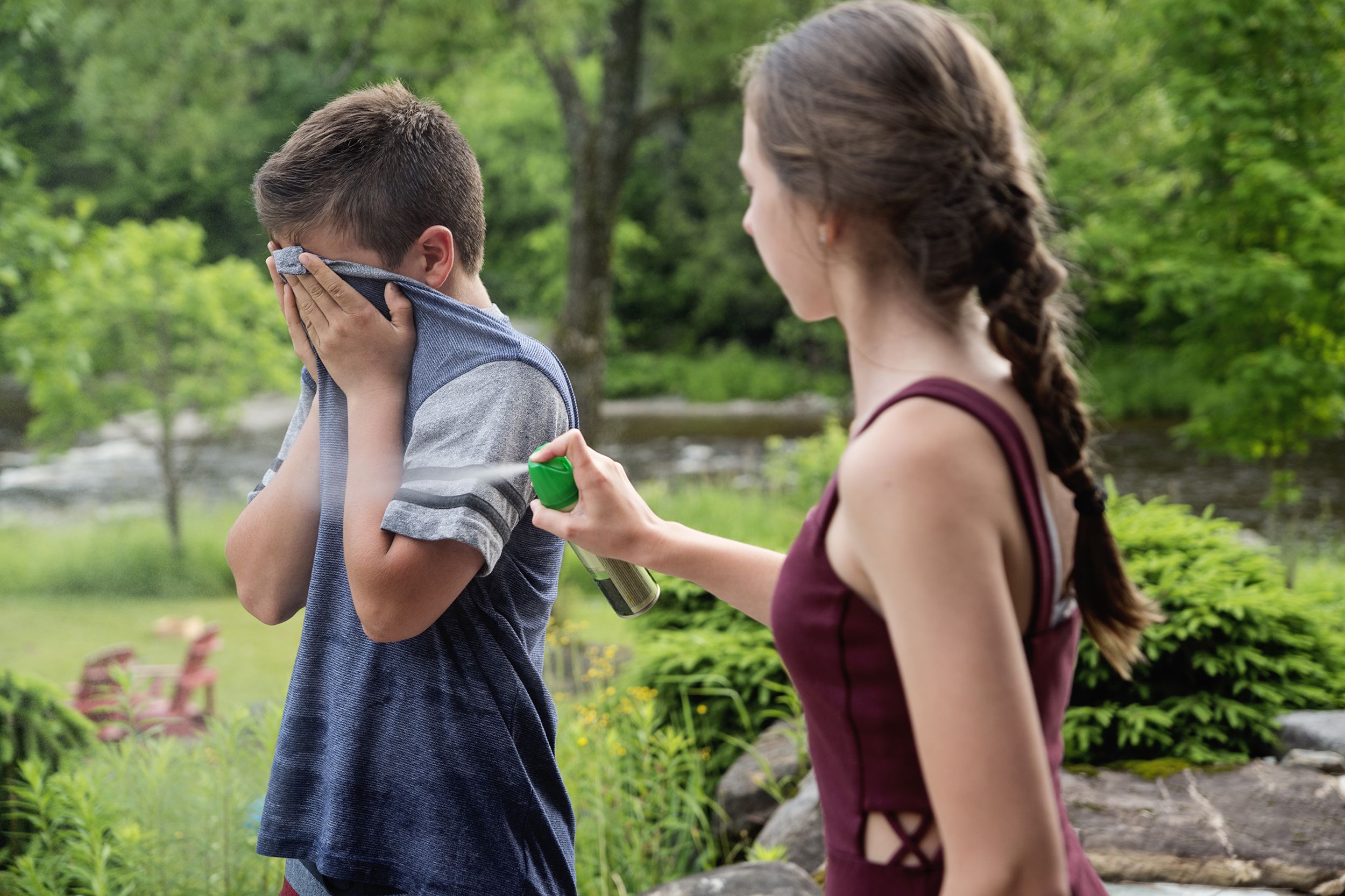 Female teen spraying insect repellent on her little male brother, girl in the right foreground spraying towards him on the left, while outside, surrounded by grass and trees on a sunny day