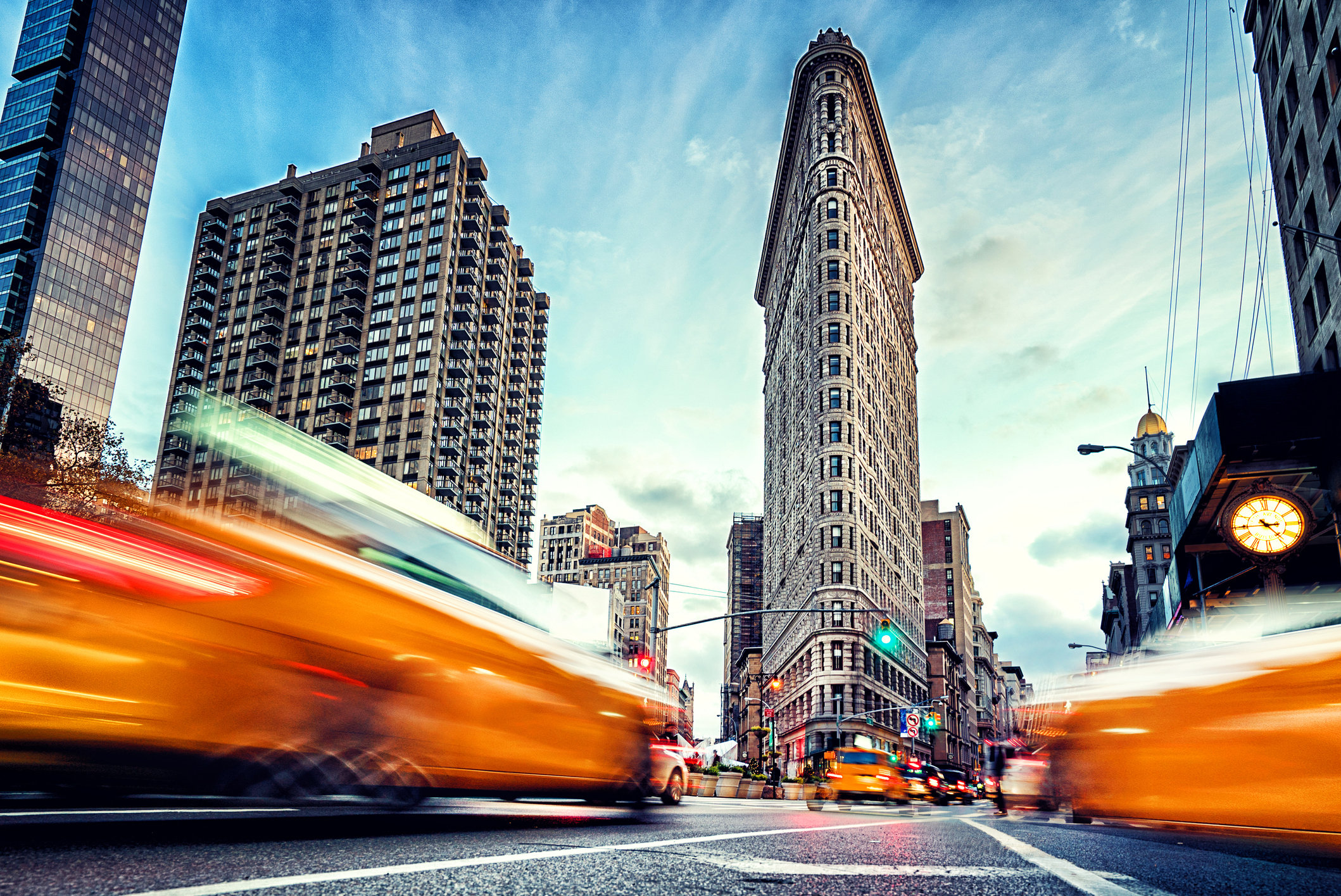 Flatiron Building, Manhattan, during sunset, yellow taxi cabs blurred in the foreground, dramatic colored sky surrounds the building