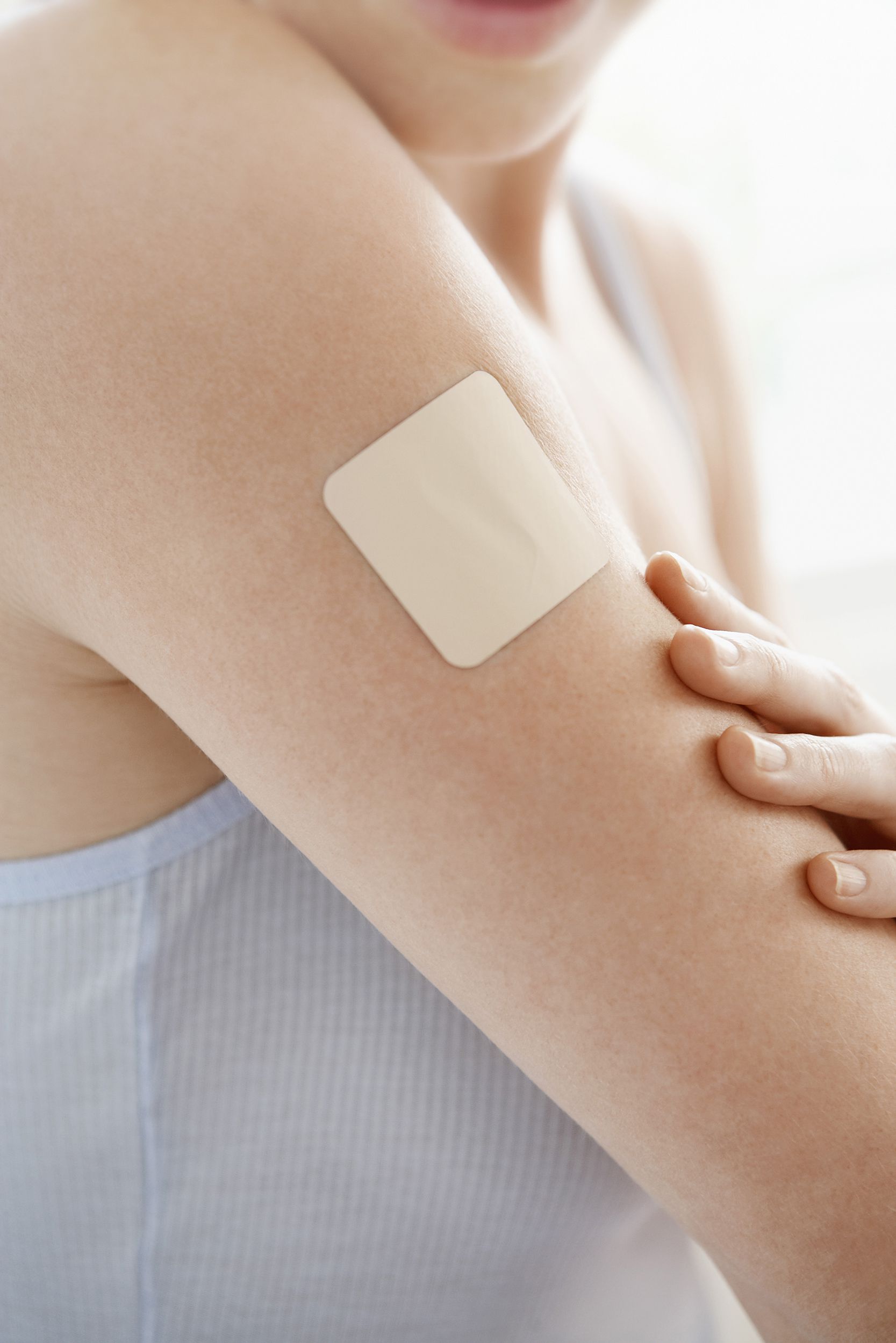 Focus on arm of woman with a beige nicotine patch, a light blue cami, blurred background of rest of her mid-section and light coming through the window