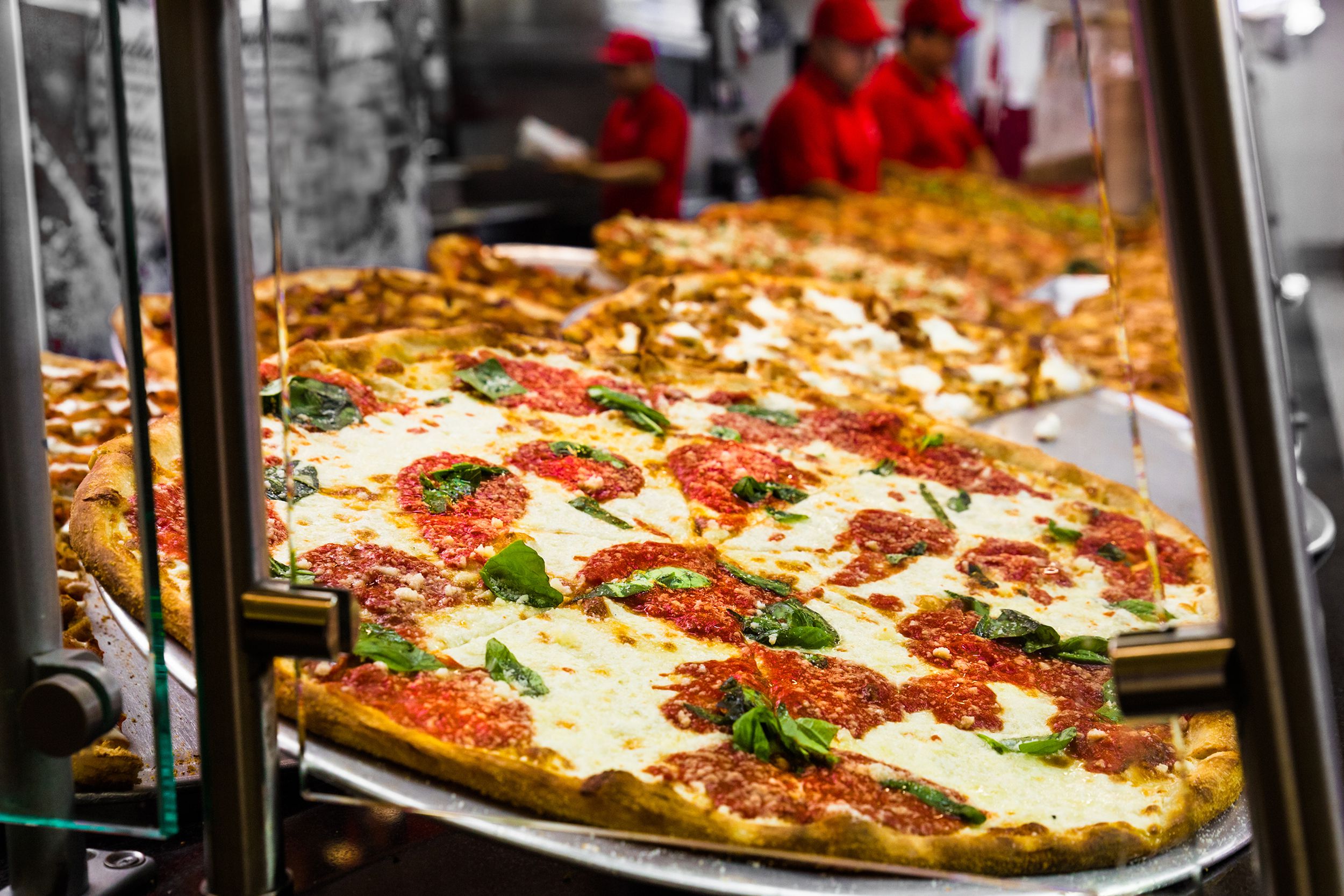 Closeup of a row of fresh Italian pizzas in a New York City pizzeria window with workers wearing red in the background