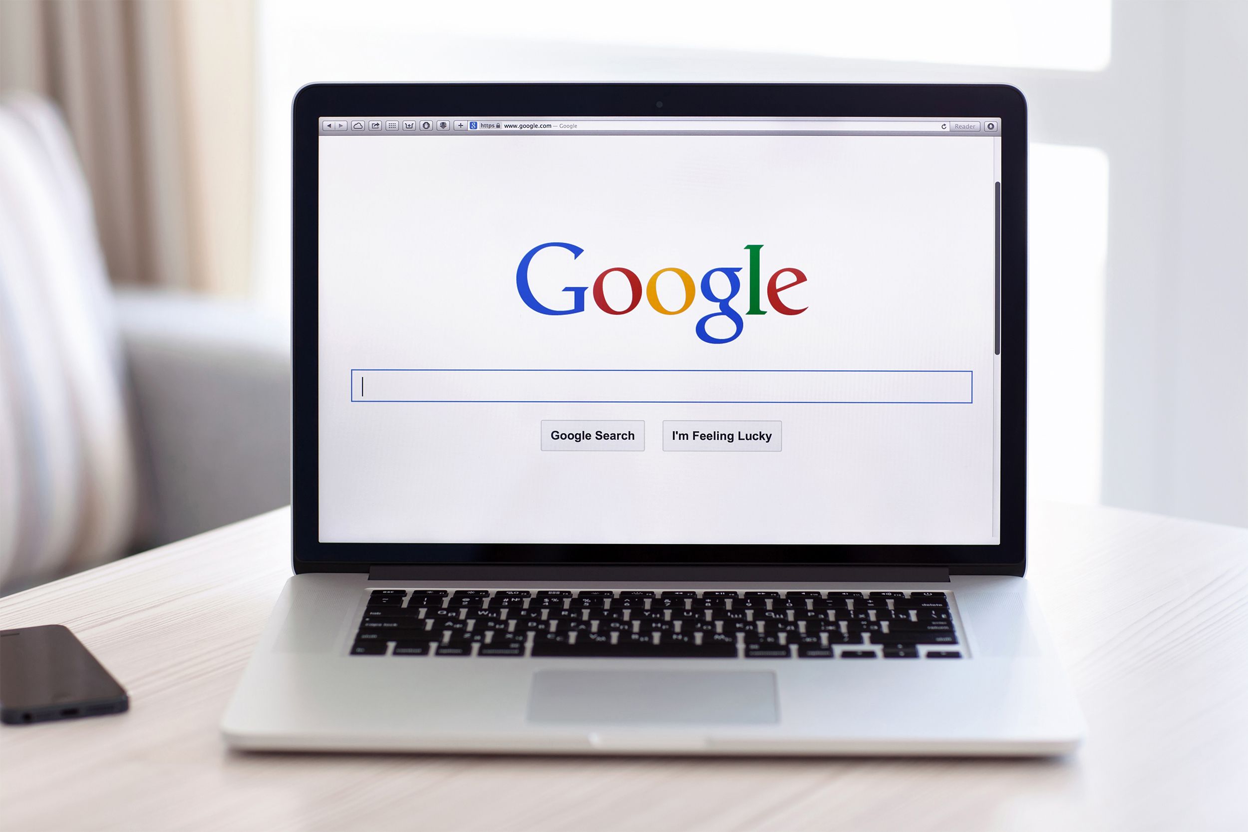 Google homepage on the safari browser on a MacBook Pro, selective focus, directly facing the camera, on a light brown table, a seat pillow, a window, and curtains blurred in the background
