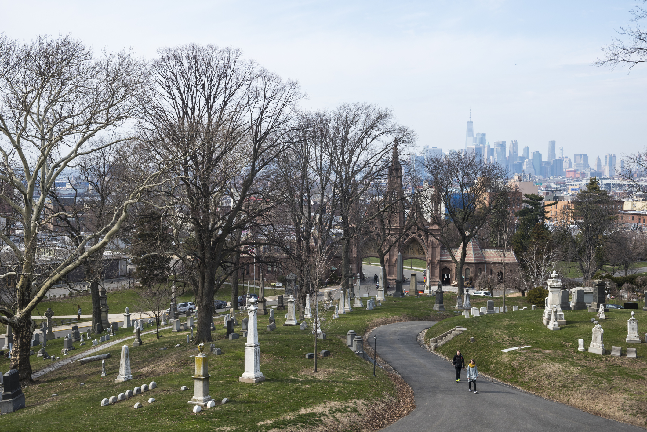 Green-Wood Cemetery, Brooklyn, during winter, two people walking along path, surrounded by numerous graves and bare trees, Manhattan skyline in the background