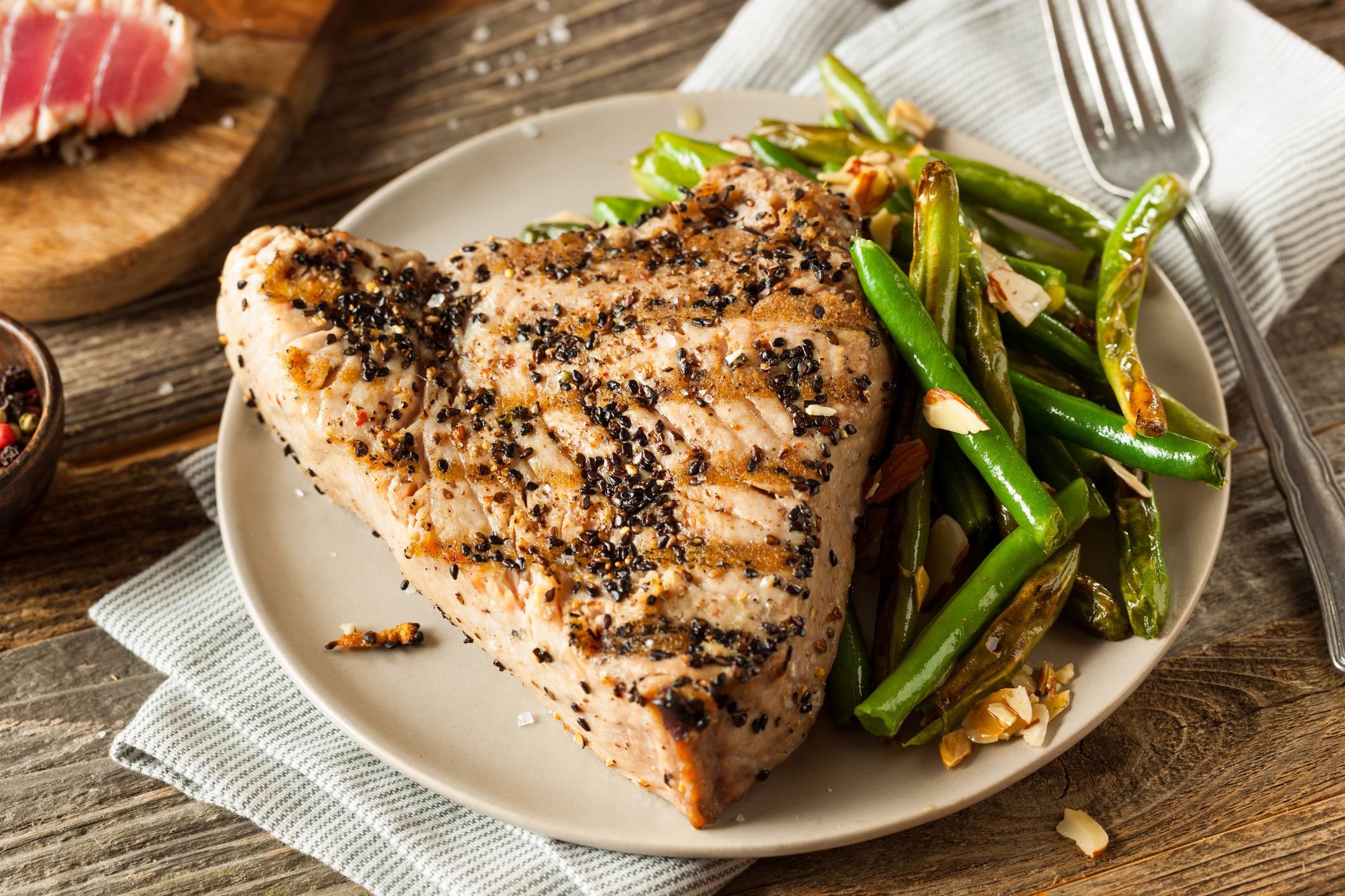 Grilled fish on a cream colored plate with a side of asparagus on a light grey napkin with a fork on a wooden table with ingredients in the background
