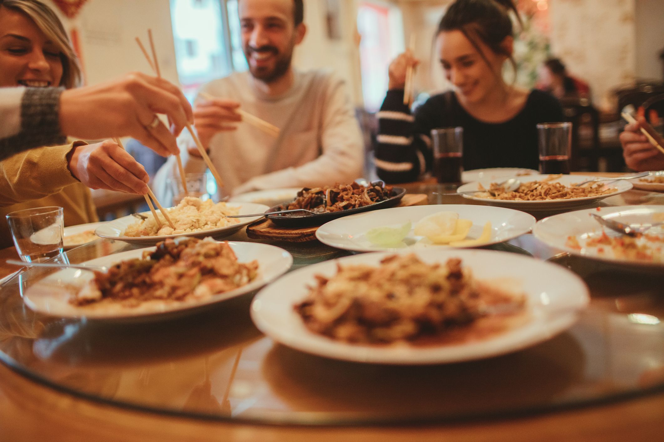Group of happy friends eating at a round table in a Chinese restaurant with focus on white plates of Chinese food with a blurred background of the restaurant