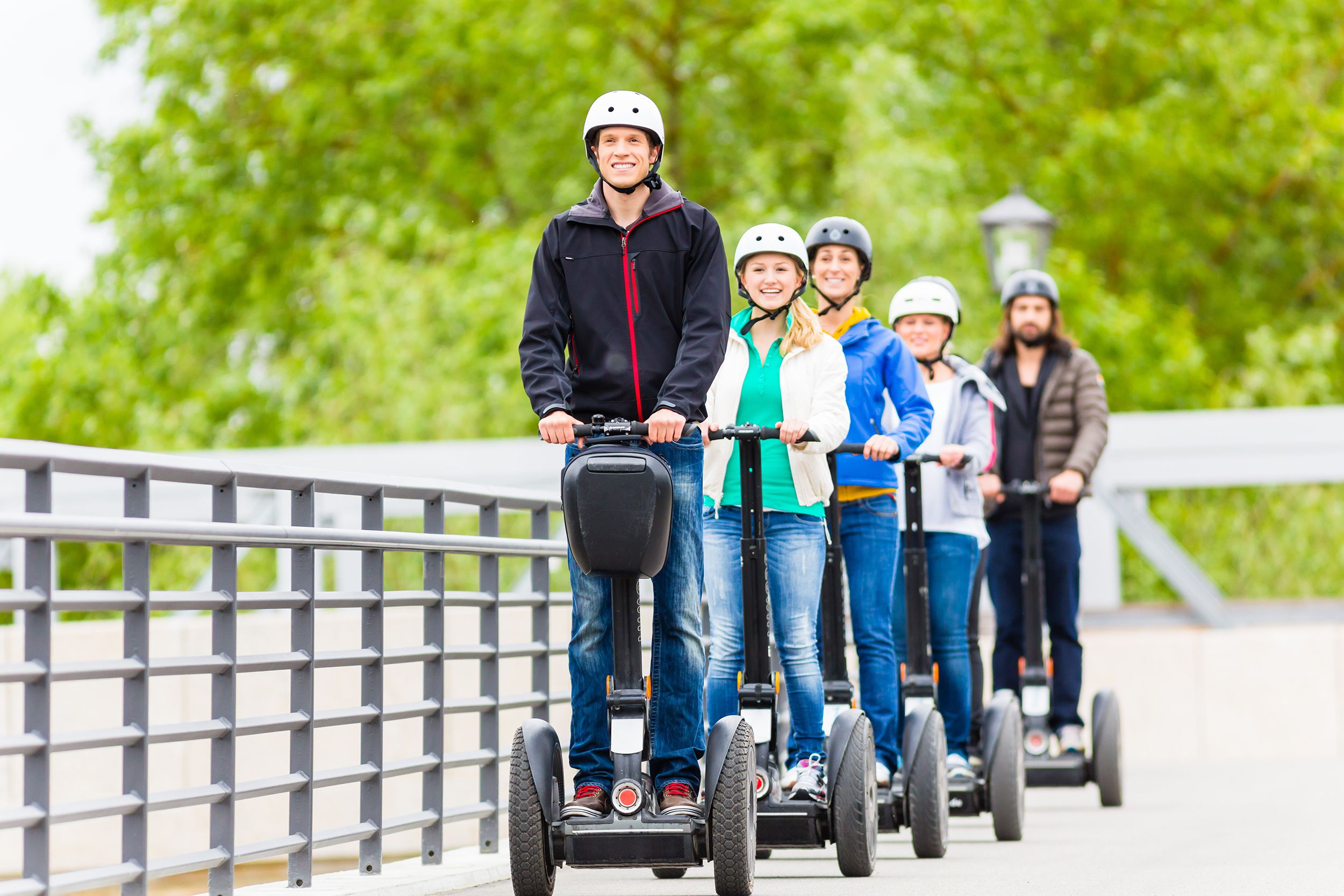 Group of six tourists all on individual Segways in a line on a sidewalk during a city tour in Germany, a chrome fence on the right, on a sunny day