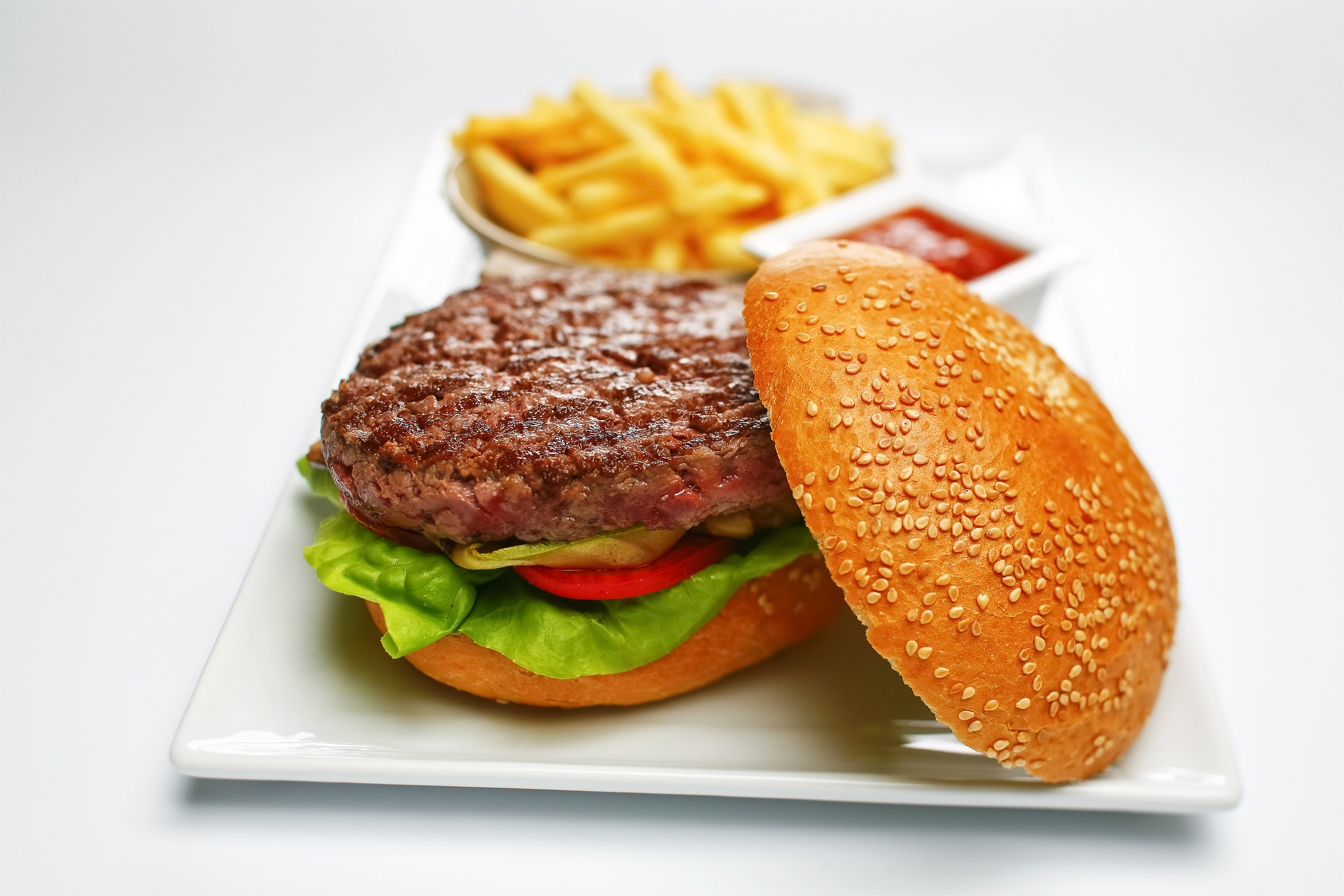 Hamburger on bottom bun with the top bun with sesame seed on the right side, lettuce and tomato on the bottom bun, on a rectangle white porcelain plate, in the foreground, fries in a stainless steel bowl and ketchup in a small square white bowl in the bac