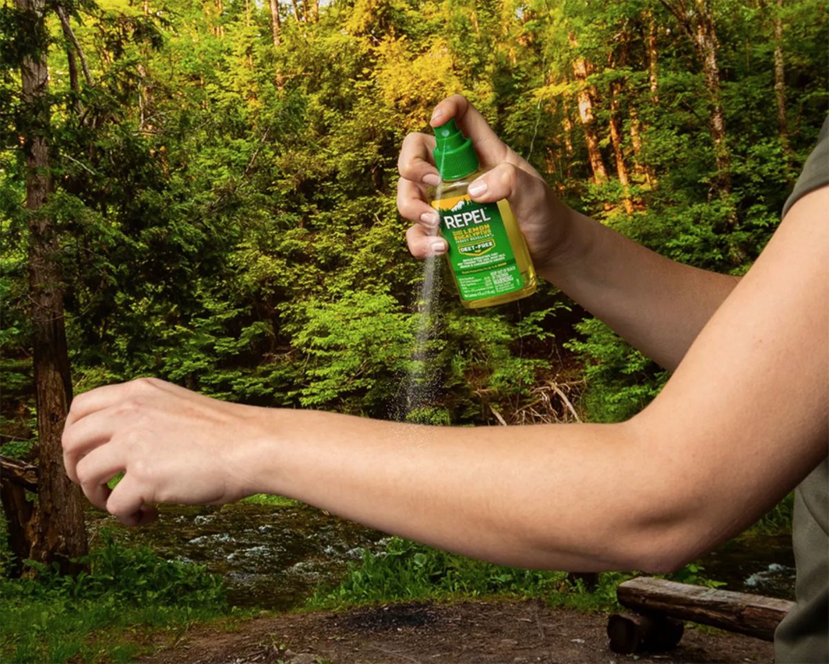 Hand holding Repel Insect Repellent spraying it on other arm, arm in the foreground, forest in the background