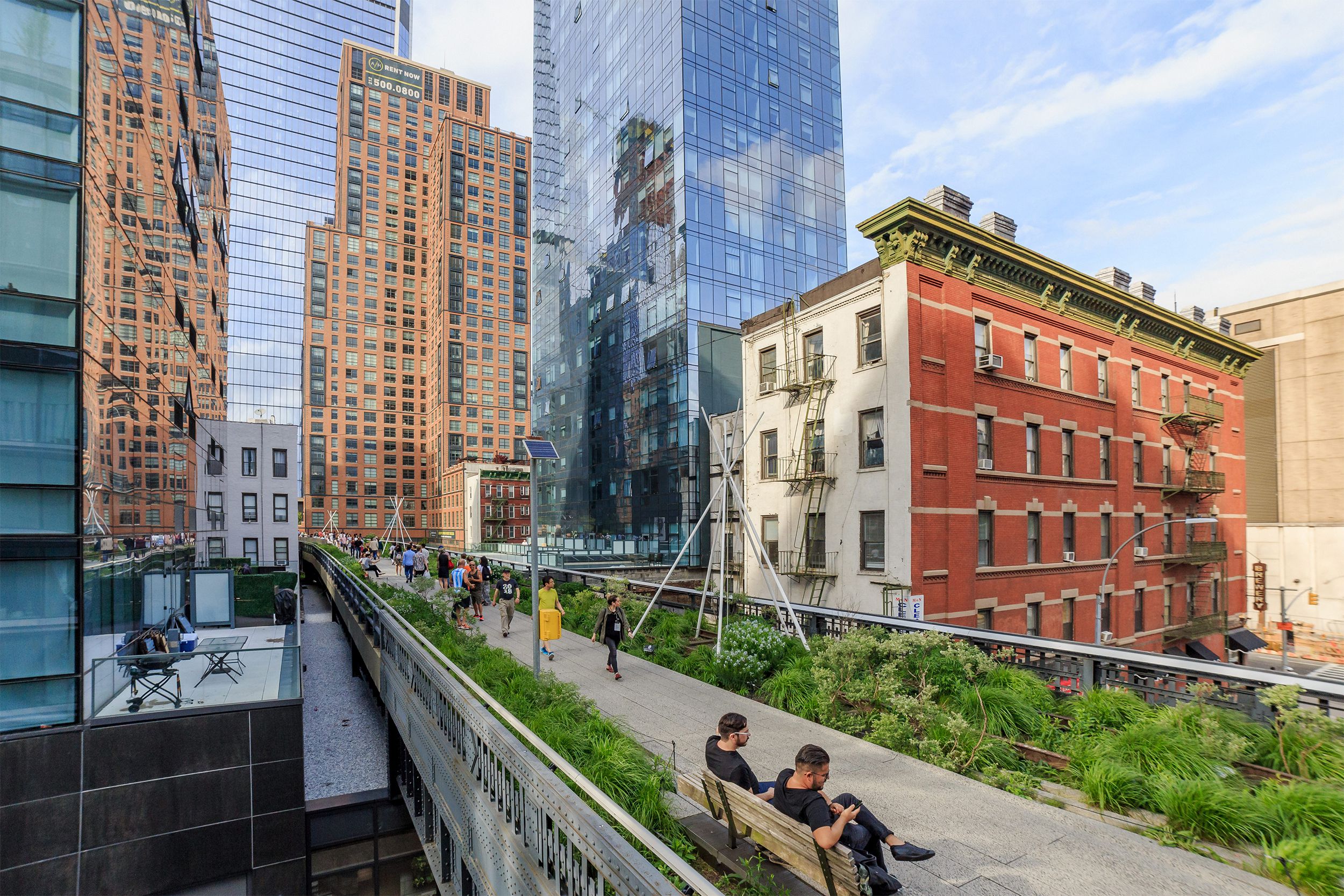 High Line Park, Manhattan, skyscrapers in the background with people walking along it, two people seated on a bench in the foreground, on a sunny day