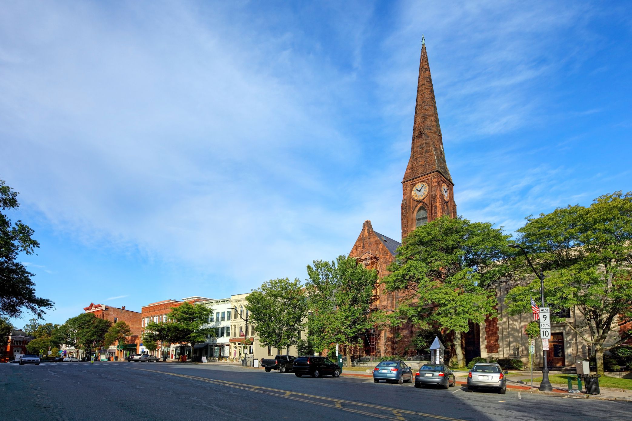Historic buildings line the street in Northampton, Massachusetts, cars parked along the parking spots, a large church, during summer, dramatic white clouds in a clear blue sky