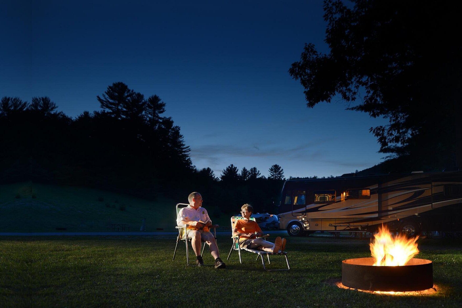 Lake George RV Park, Lake George, New York, a couple sitting together at a fire pit in a lot with RV in the background during late evening