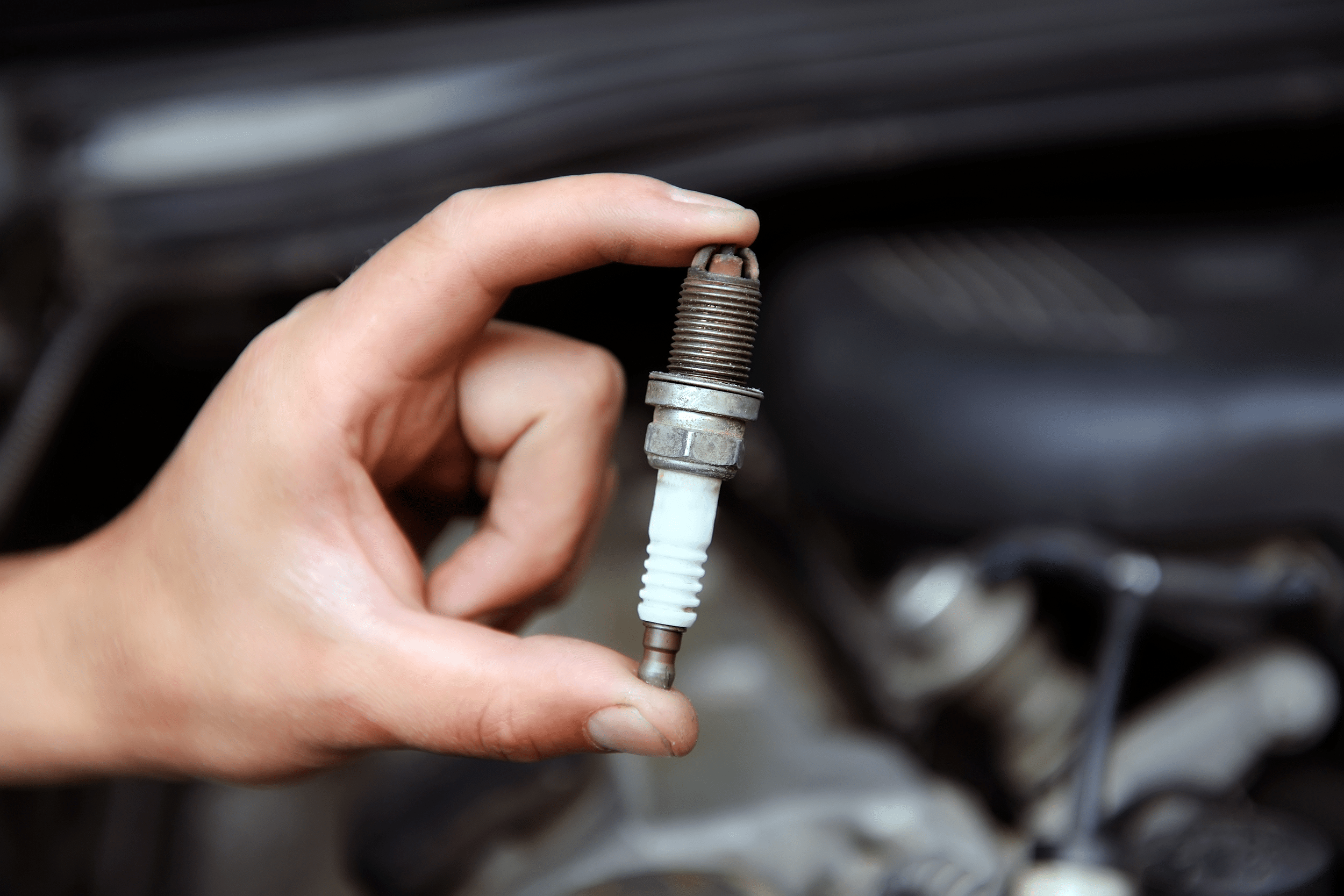 Left hand holding a used spark plug with thumb and pointer finger, over a blurred image of a car engine