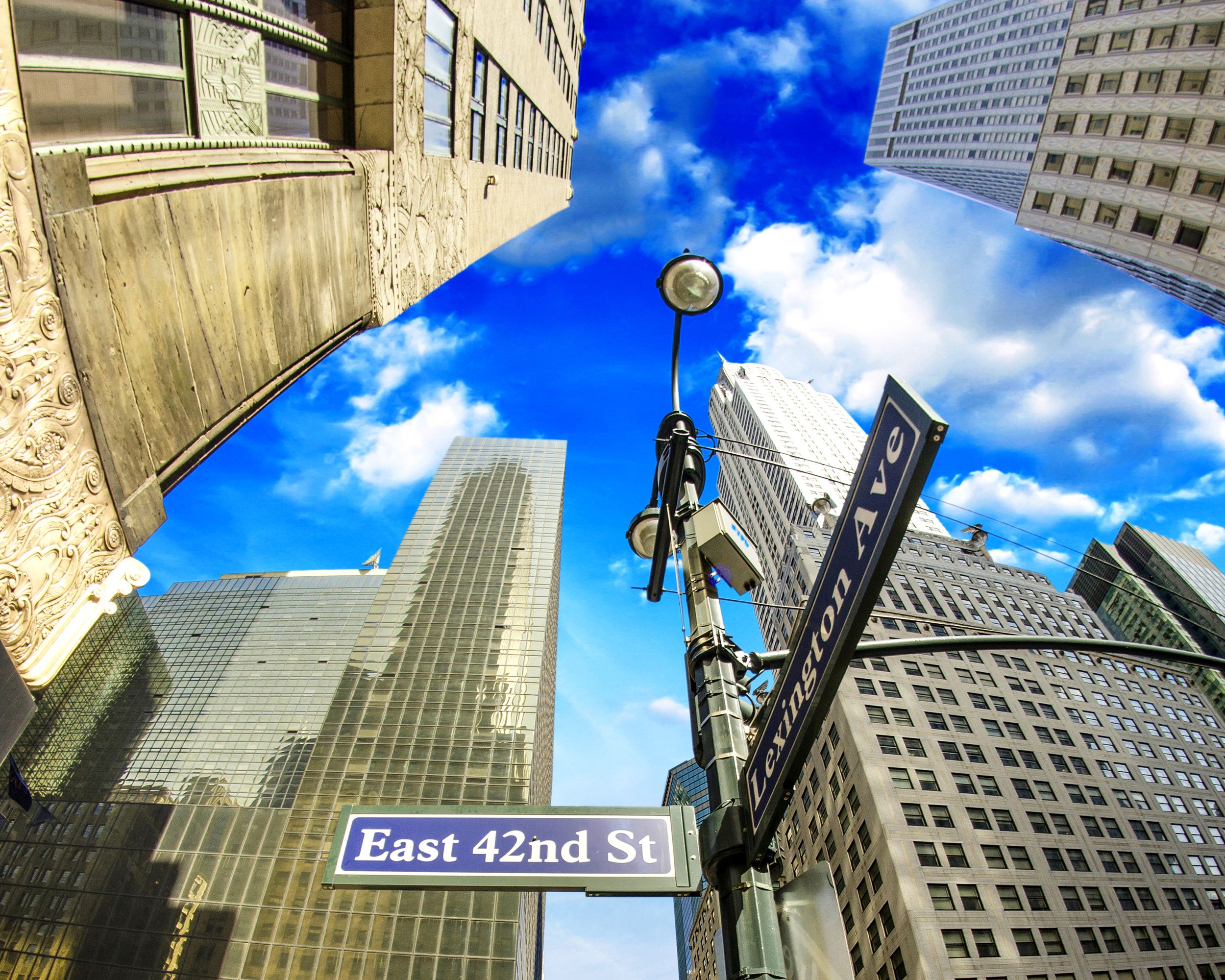 Looking up towards the sky with skyscrapers around in Midtown, Manhattan, two street signs in the foreground, East 42nd and Lexington Ave., on a bright sunny day with white clouds