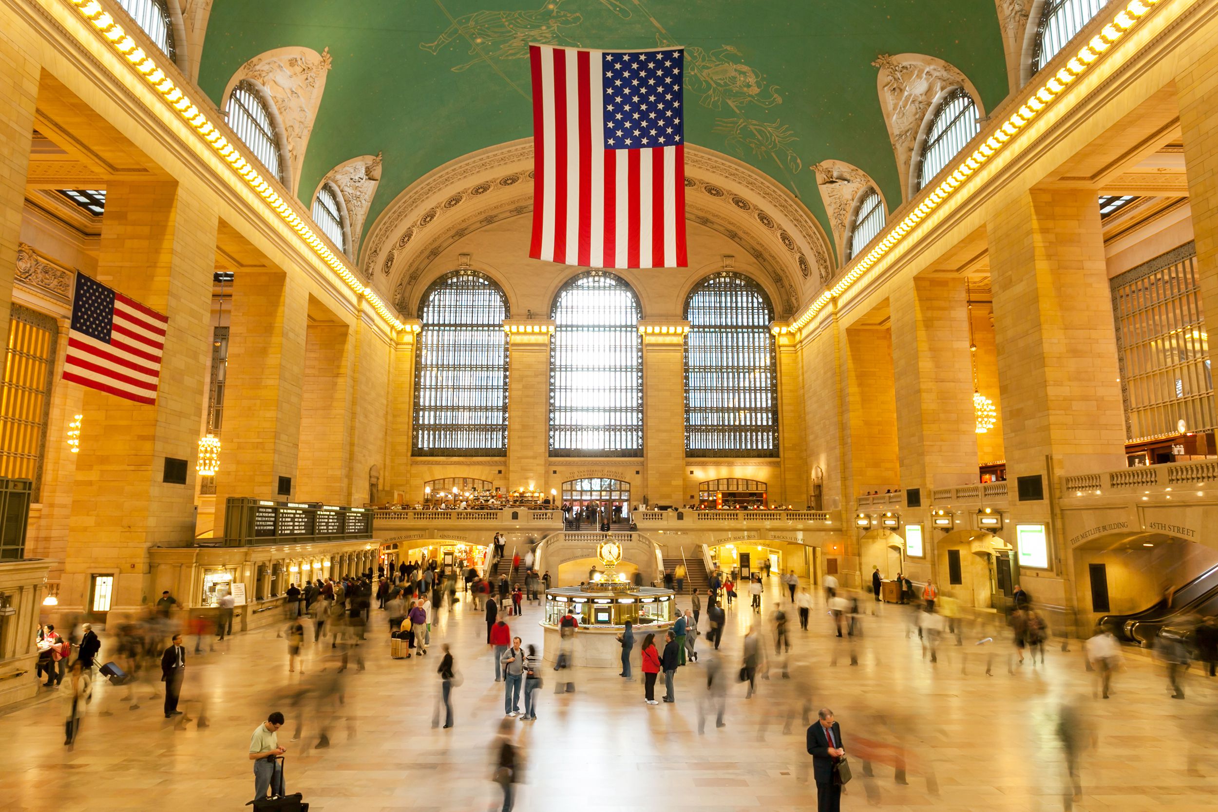 Main lobby at the Grand Central Terminal, Manhattan, American flag hanging in the center, people in motion, some blurred, light is coming through the large windows