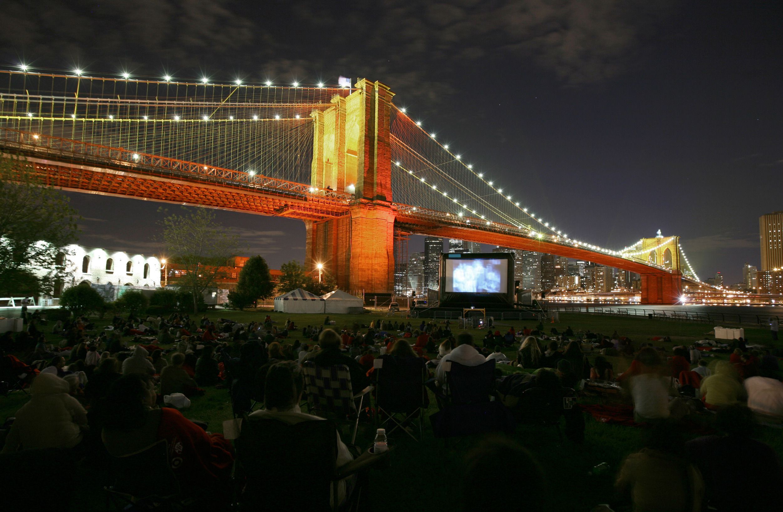 Crowd of people watching an outdoor movie at the Brooklyn Bridge Park, Brooklyn during night, the Brooklyn Bridge behind it, lower Manhattan lit-up in the background