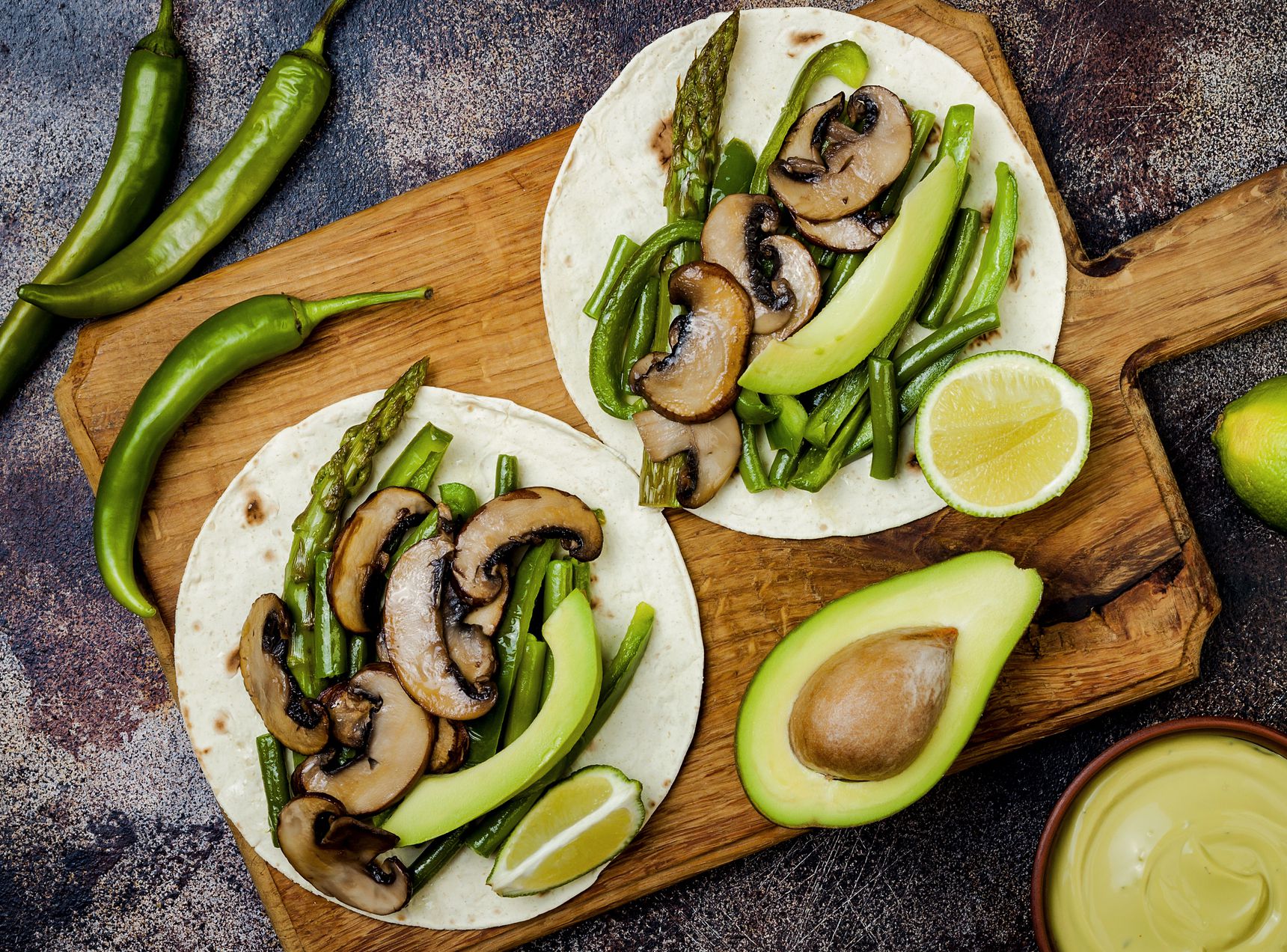 Two portobello mushroom tacos on a wooden server with chilis, avocados, and limes in the background