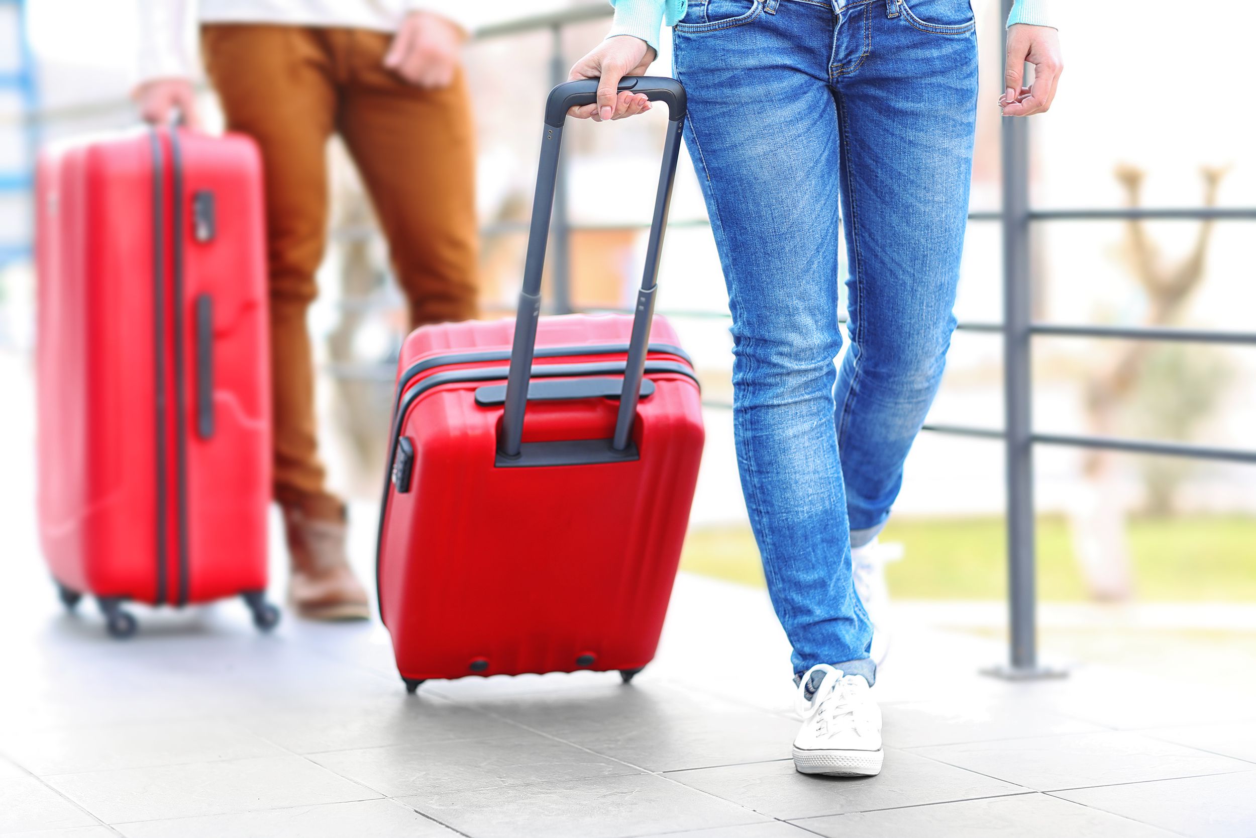 Red roller carry-on luggage being rolled by a woman, legs of woman wearing jeans, man with larger roller red luggage blurred in the background, outside