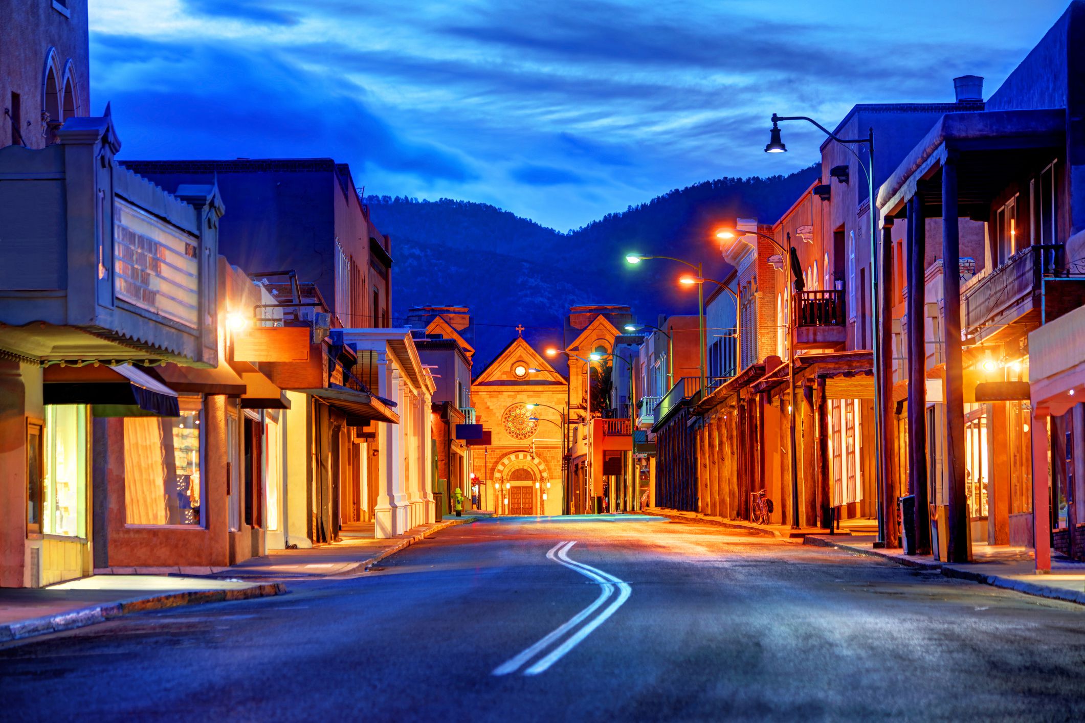 Road with shops on both sides in downtown Santa Fe, New Mexico, during evening, lights line the road, a church in the middle in the distance, road is in the foreground, blue mountains in the background