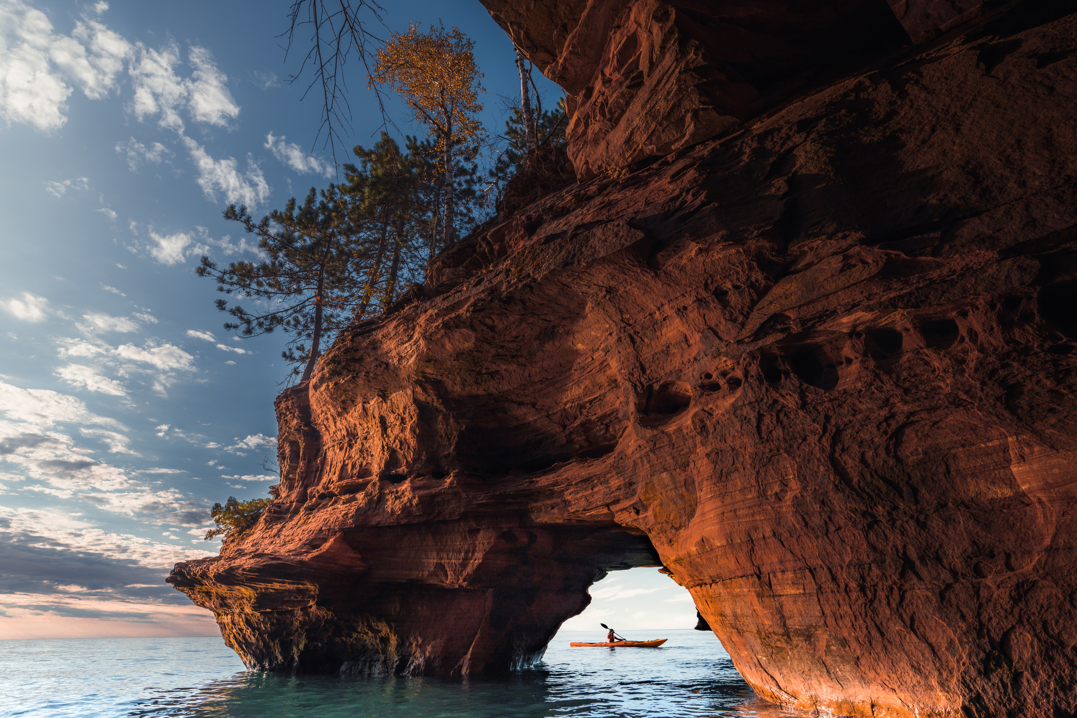A lone kayaker through an opening in a sea cave, Apostle Islands National Lakeshore, Wisconsin with an expansive cliff, early sunset in the background