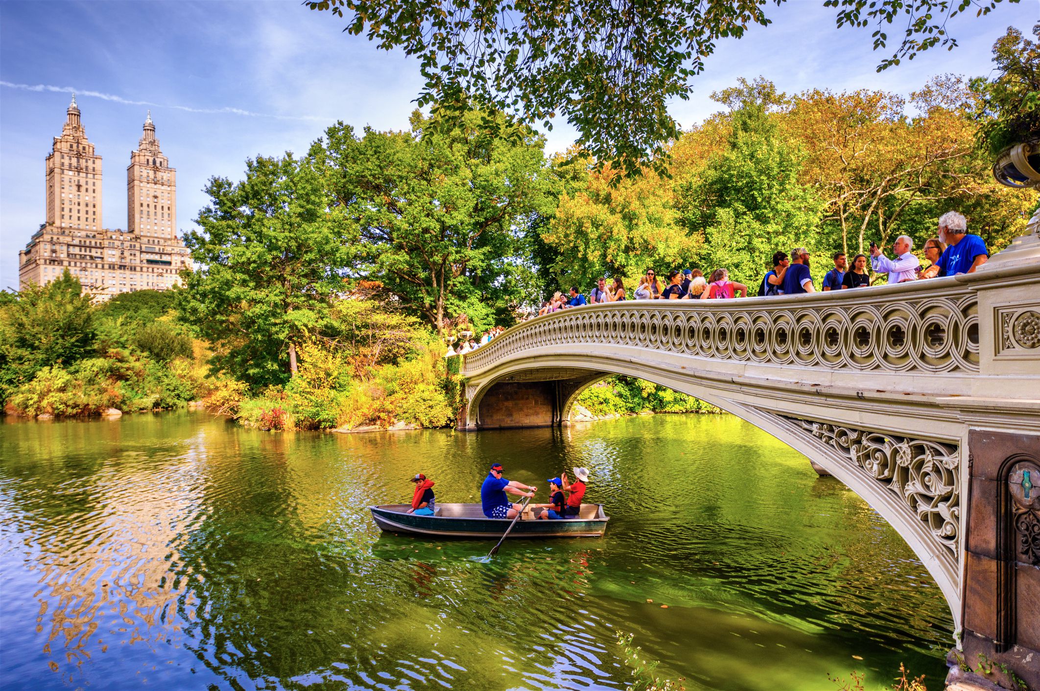Three people in a boat under Bow Bridge, Central Park, Manhattan, people walking along the bridge, during early autumn, trees and plants in the background