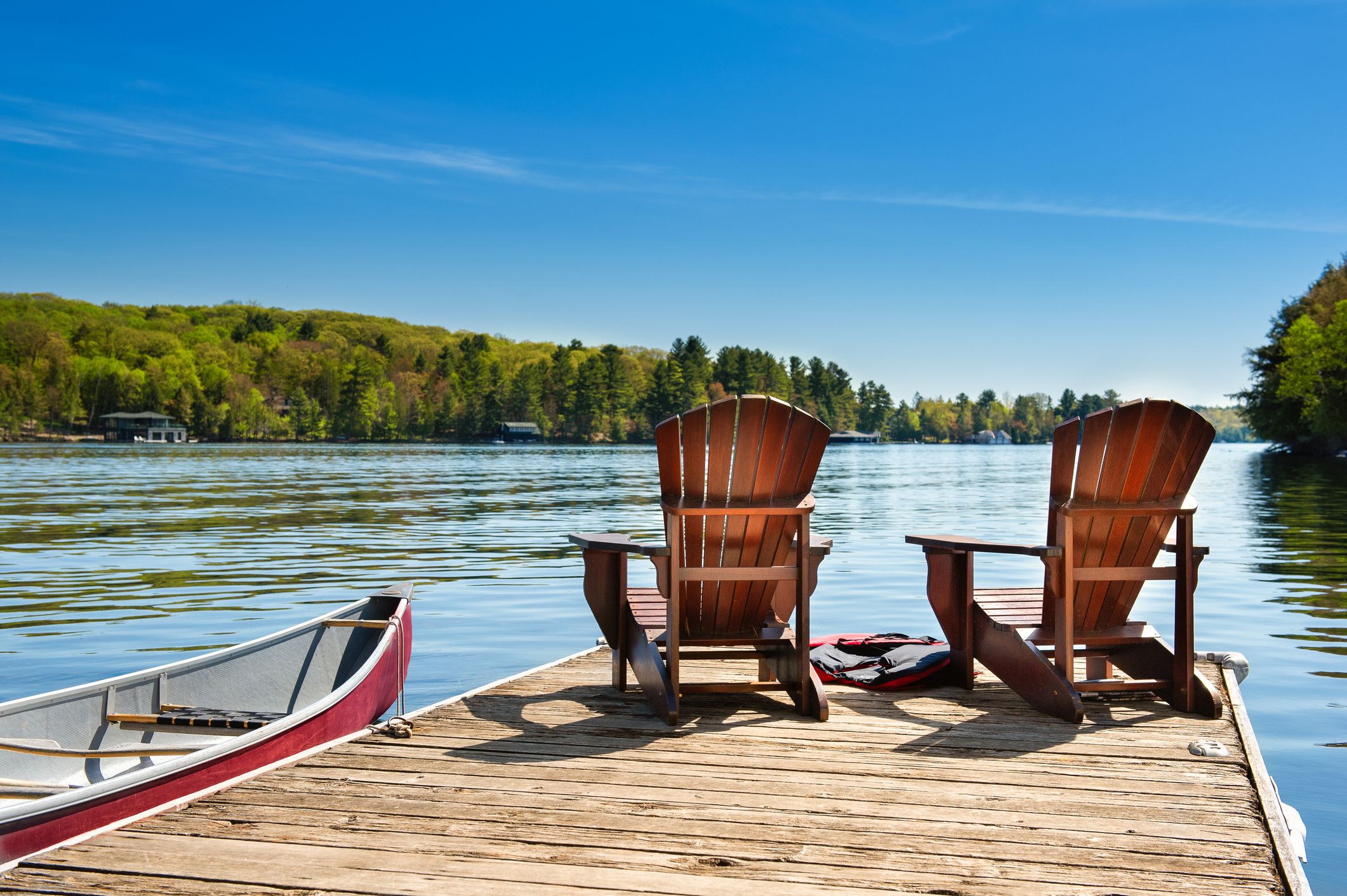Two Adirondack chairs on a wooden dock overlooking the blue water of a lake in Muskoka, Ontario, Canada, red canoe is tied to the pier and life jackets are visible near the chairs on a bright sunny day in summer