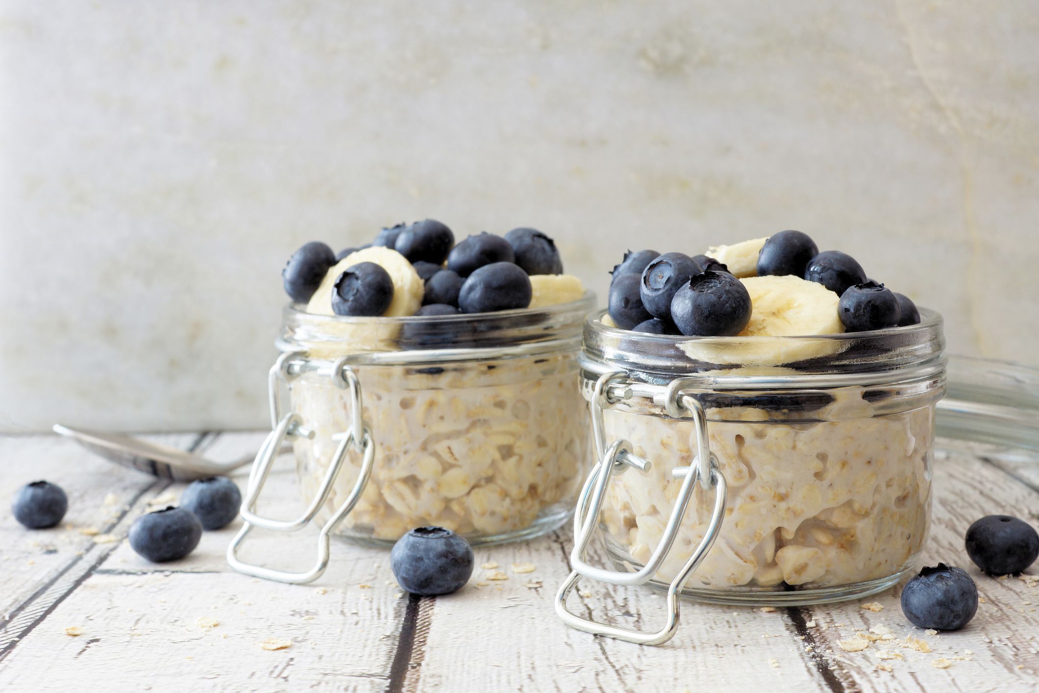 Two glass jars of overnight oats with blueberries and bananas on a white wooden table with a spoon and blueberries
