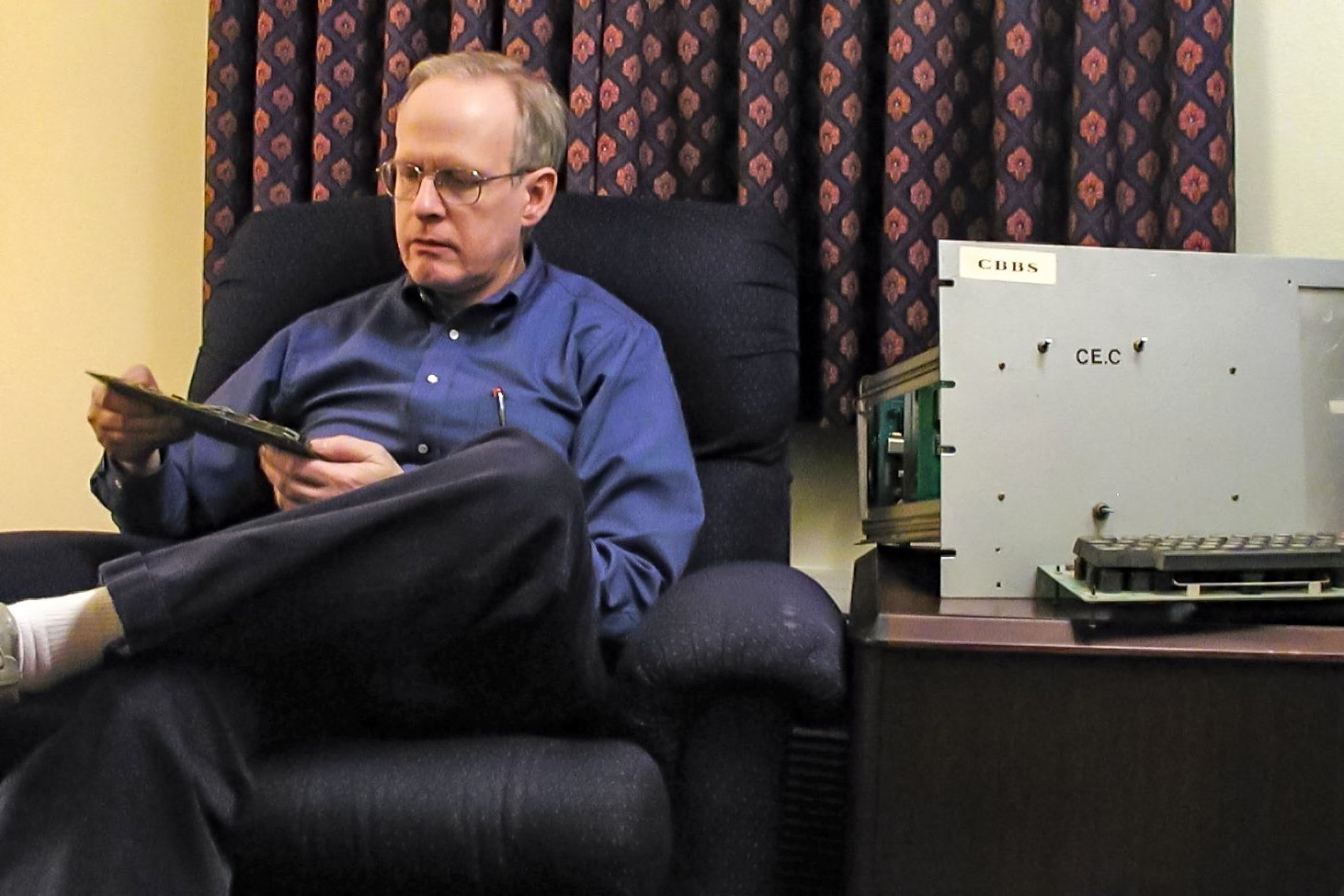 Ward Christensen holds an expansion card from the Original CBBS S-100 Host Machine, which is on the right, he is sitting on the left, beige wall and red and black curtains in the background