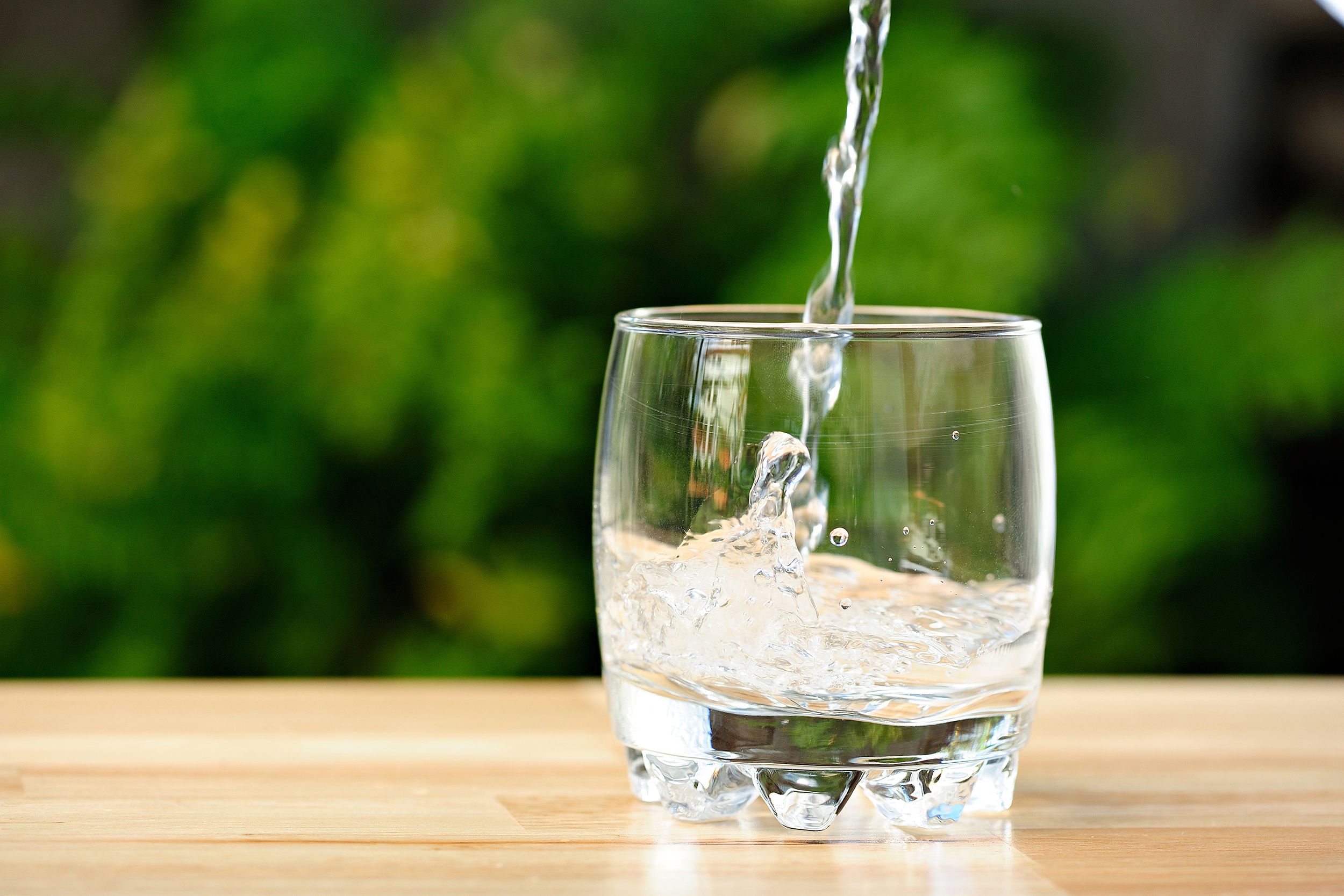 Water being poured into a whisky glass on a light wooden table, slightly on the right, greenery blurred in the background