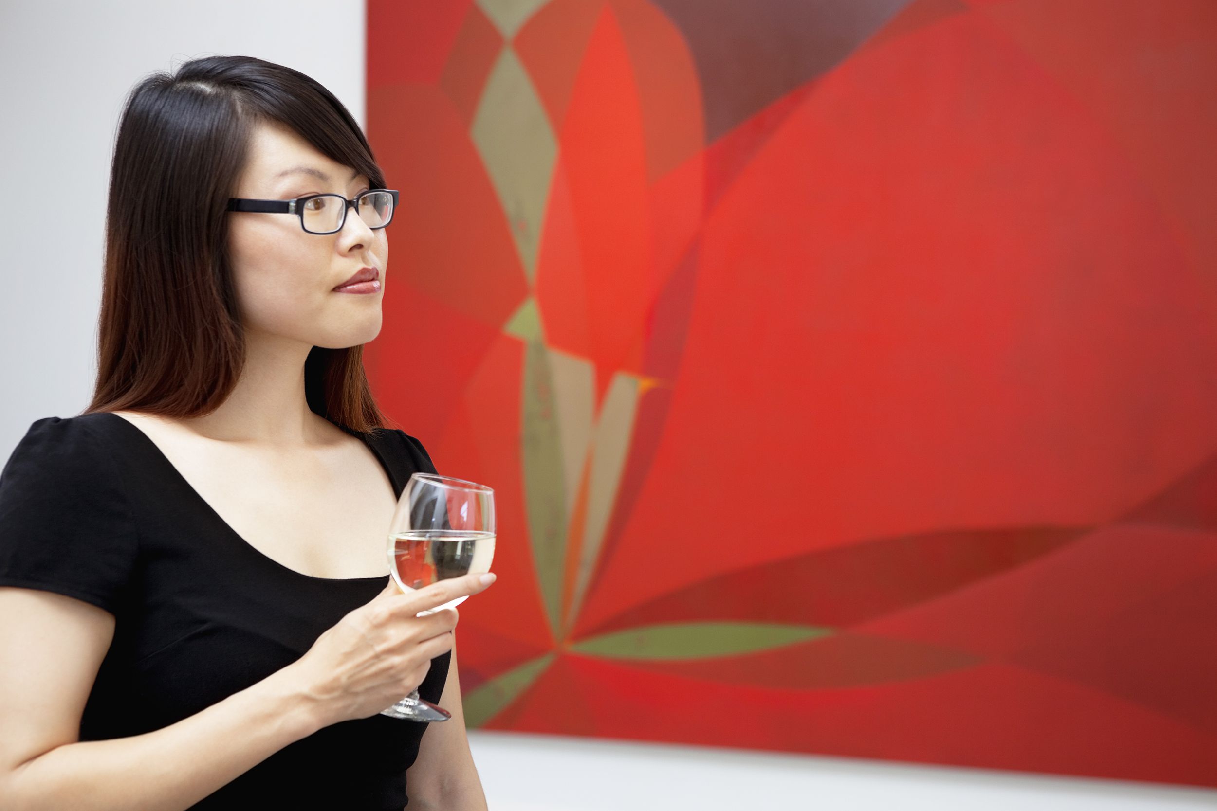 Young woman on the left, holding a glass of white wine, in a black dress, looking at a red wall painting hanging in an art gallery