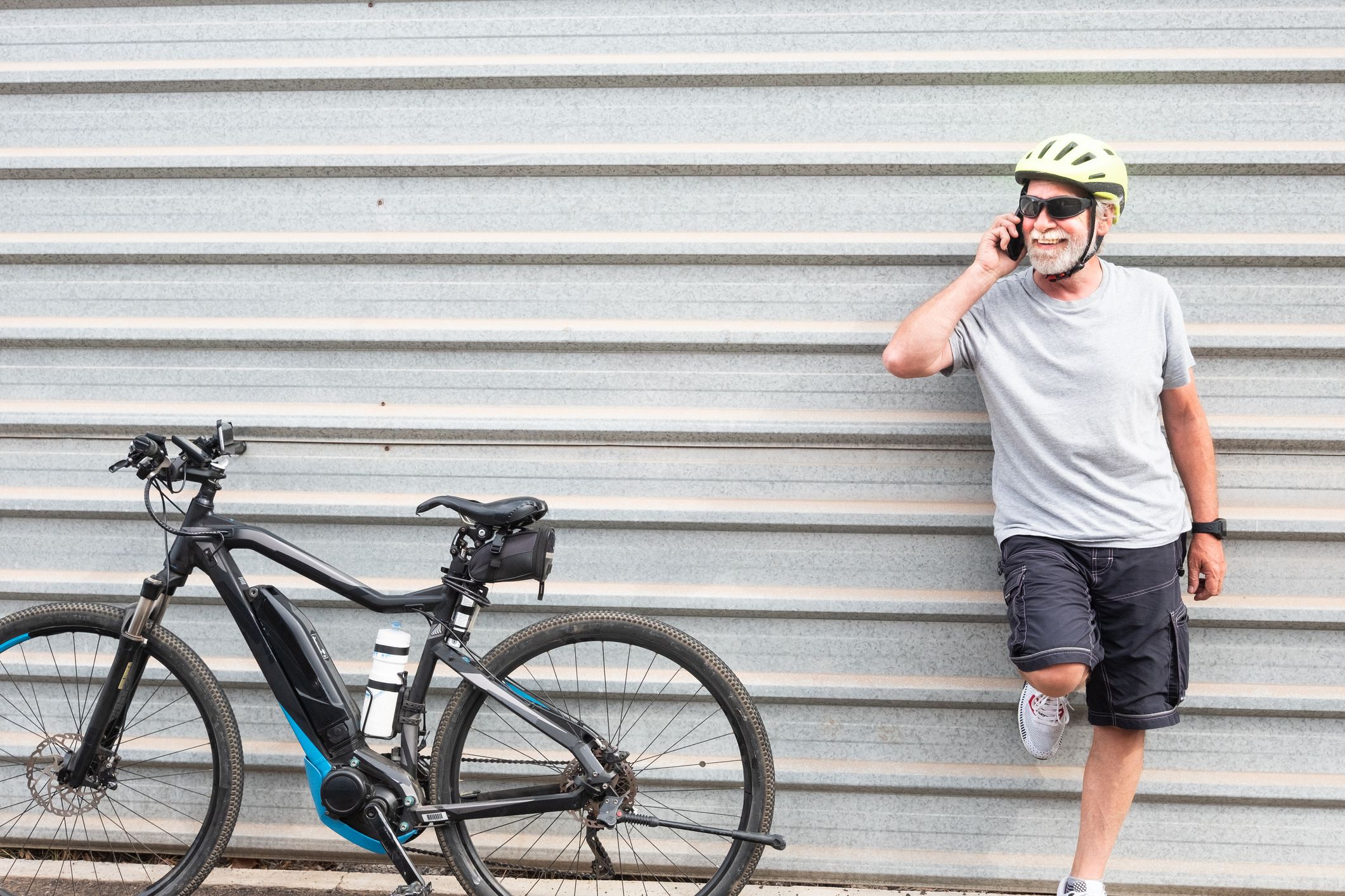 Happy senior man standing talking at mobile phone. Metal wall in background. One caucasian people. Using e bike with helmet