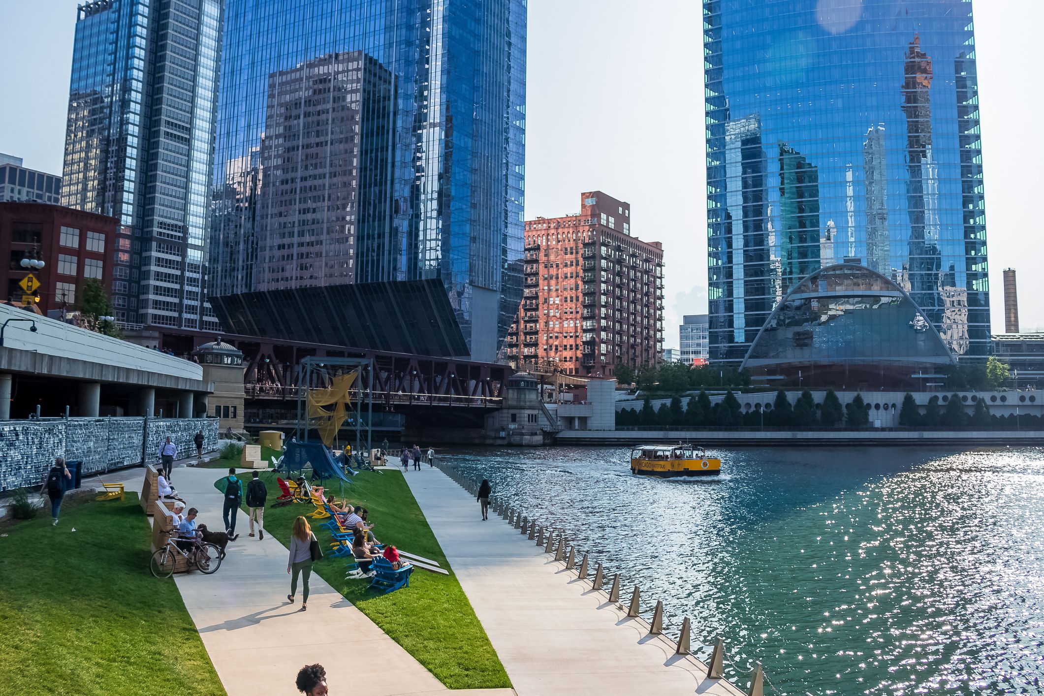 Chicago, IL / USA - 7/23/19: Sunny summer afternoon along Chicago River riverwalk where people walk and relax while water taxi crosses river