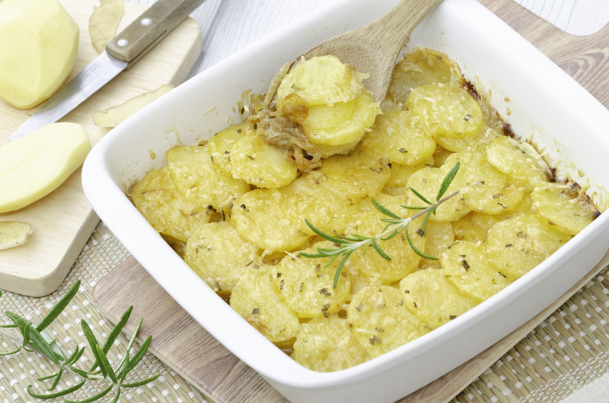 Scalloped potatoes gratin with caramelized onions and Gruyere and Parmesan cheese, sprinkling of rosemary in a square white gratin dish with peeled raw potatoes in background