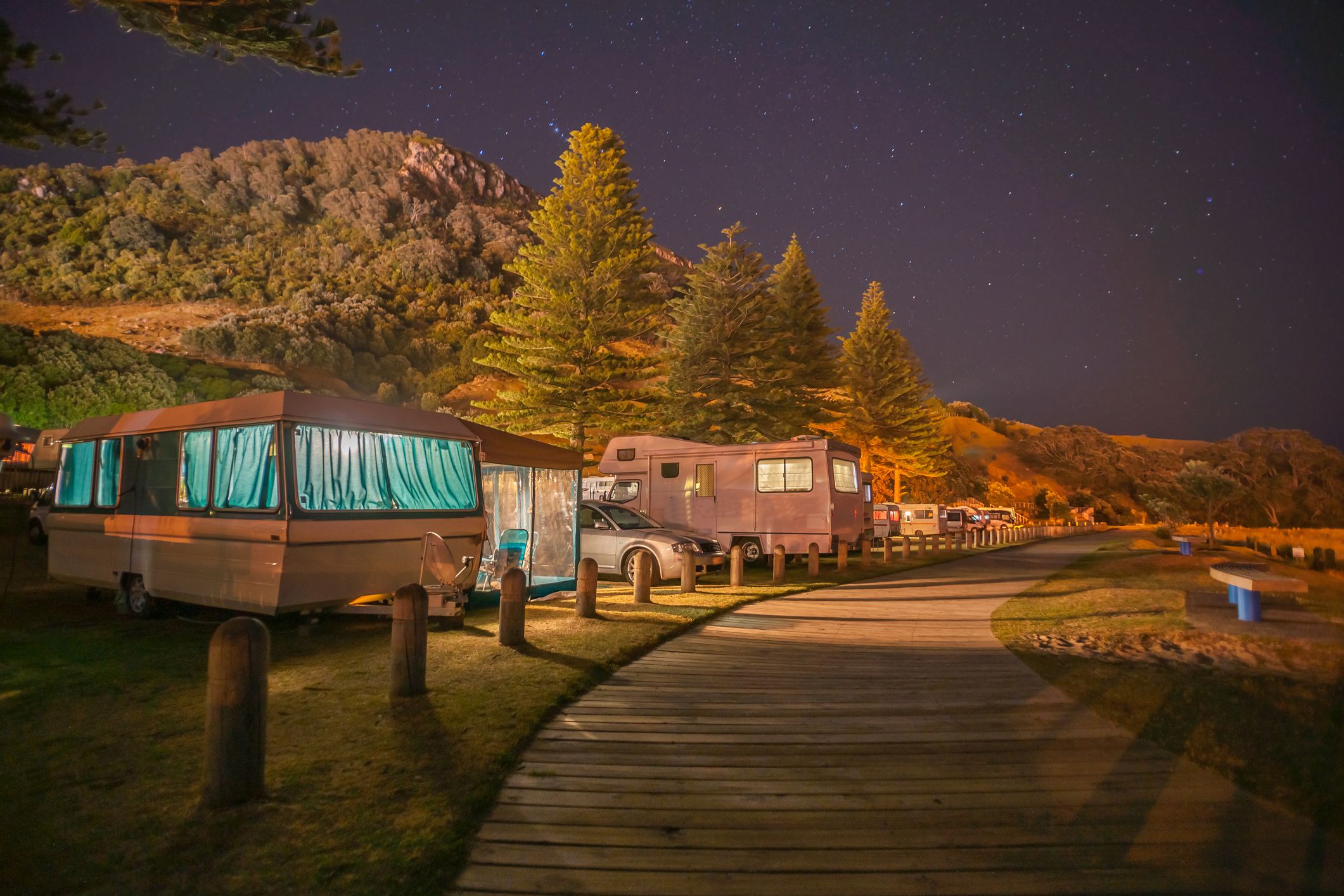 Caravans and mobile homes of holidaymakers under stary sky along wooden walkway to base of Mount Maunganui New Zealand.
