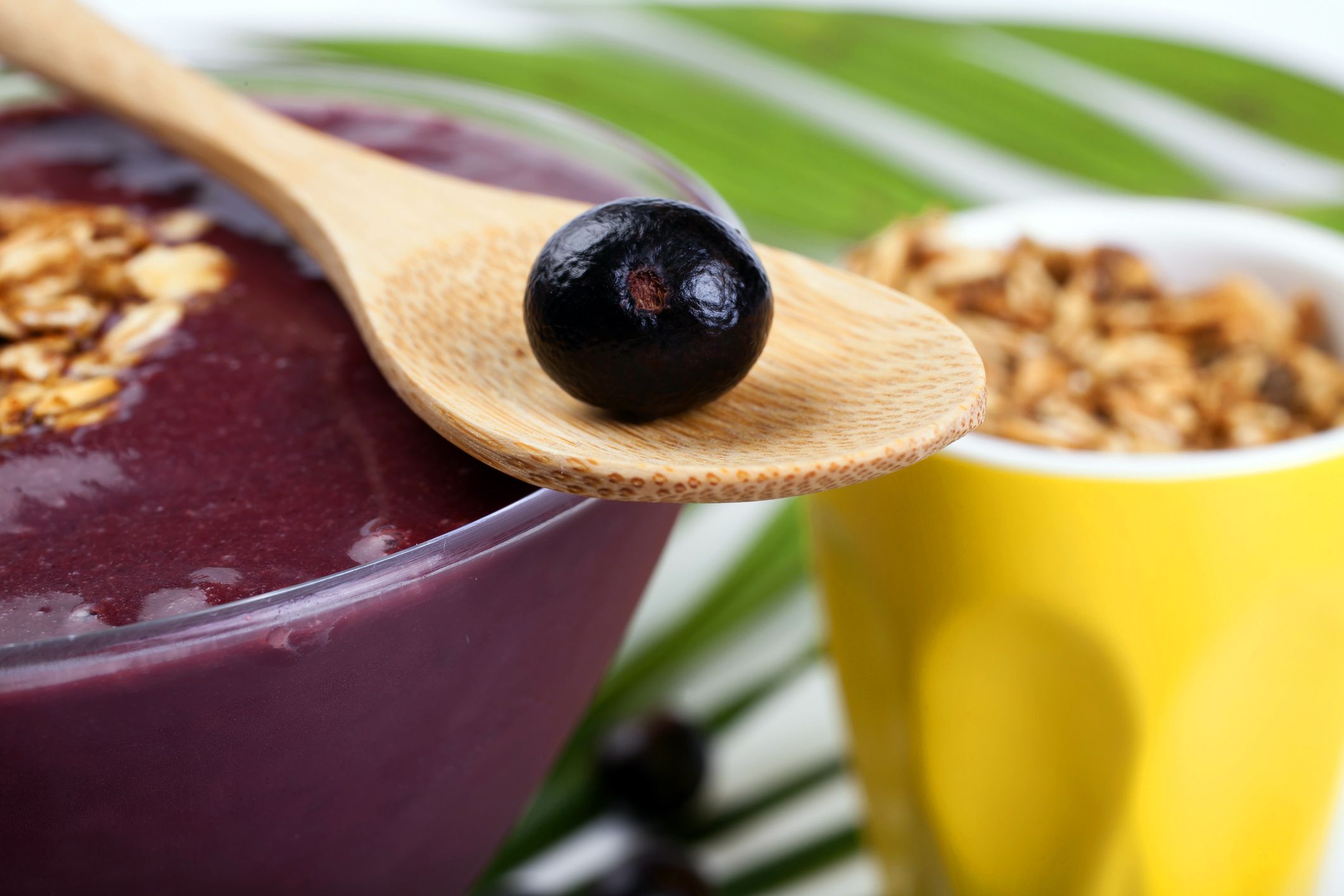 acai bowl with granola on top, glass of granola in background, wooden spoon with acai berry in foreground