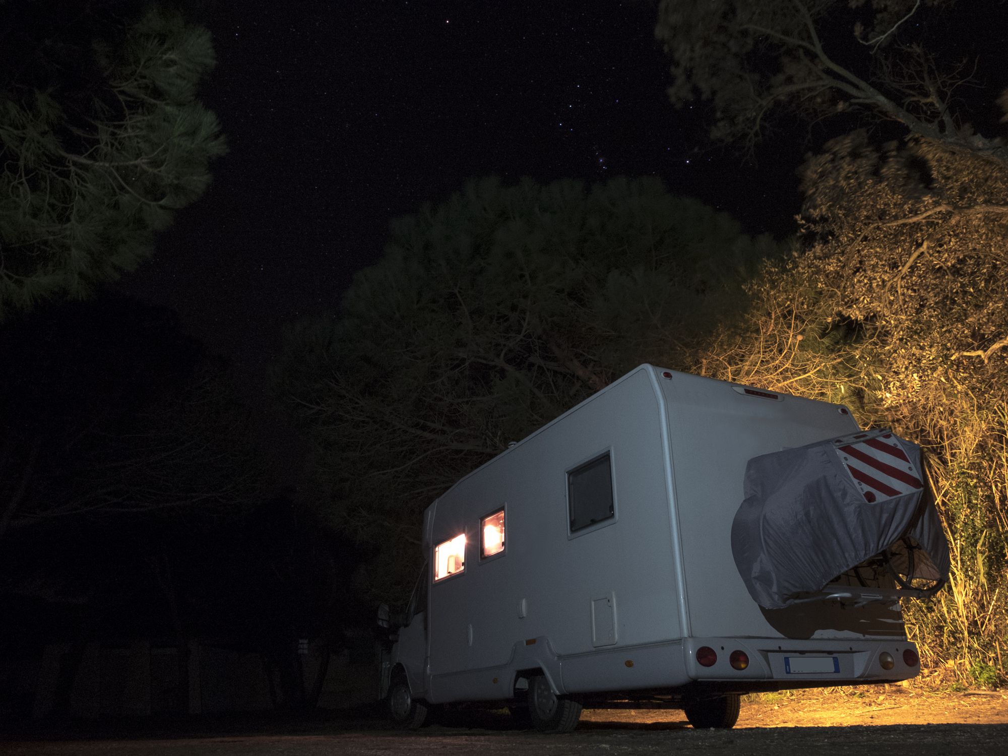 White camper parked in a pine forest, on the sand, under a starry sky, lights on inside the camper and out
