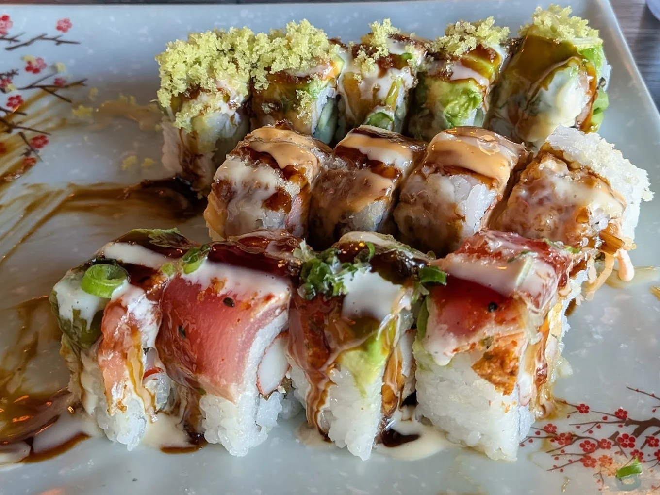 Close-up of assorted sushi rolls topped with colorful fish, avocado, sauces, and crunchy flakes, arranged on a decorative plate with a floral design.