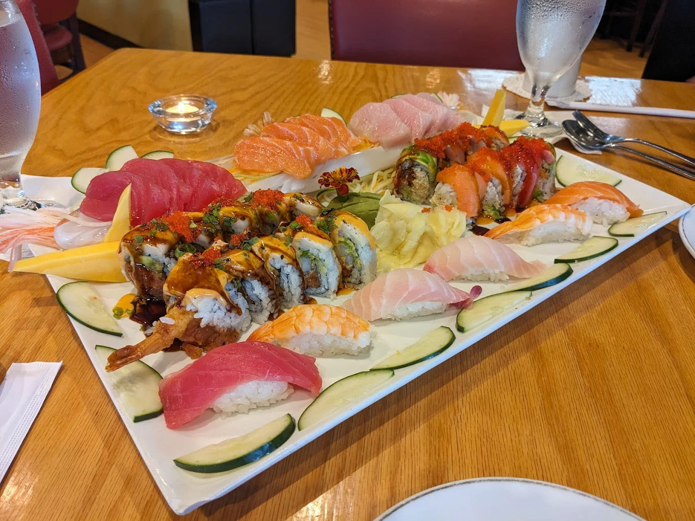 A large white platter with assorted sushi and sashimi, including colorful rolls, nigiri with shrimp, tuna, and salmon, garnished with cucumber slices, wasabi, and ginger, on a wooden table with water glasses.