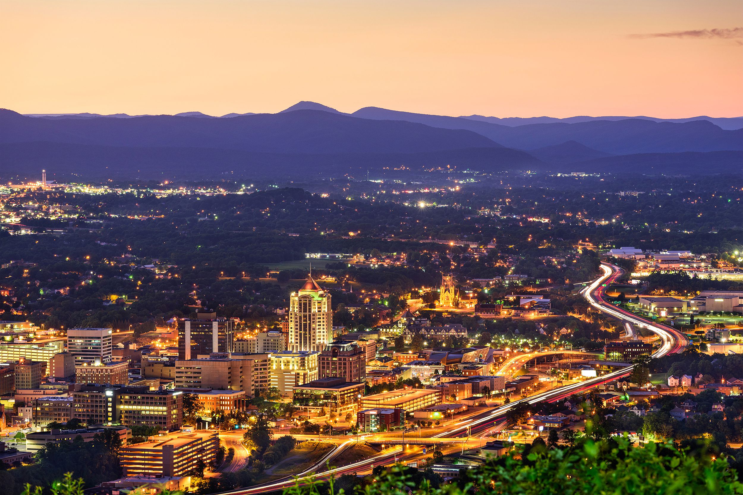 Aerial of downtown skyline of Roanoke, Virginia, during dawn, lights in buildings and along roads, blue mountains in the background against a yellow-orange sky