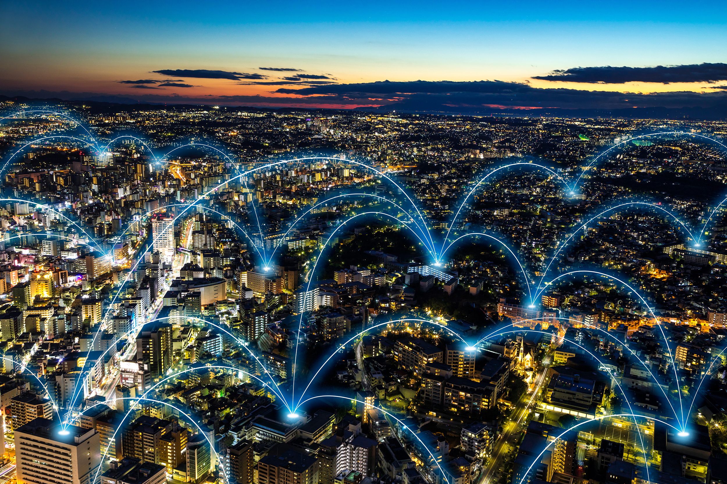 Blue 5G cellular data network over a modern city well-lit during late sunset, with a completed sunset in the background and a dark blue sky looming over it